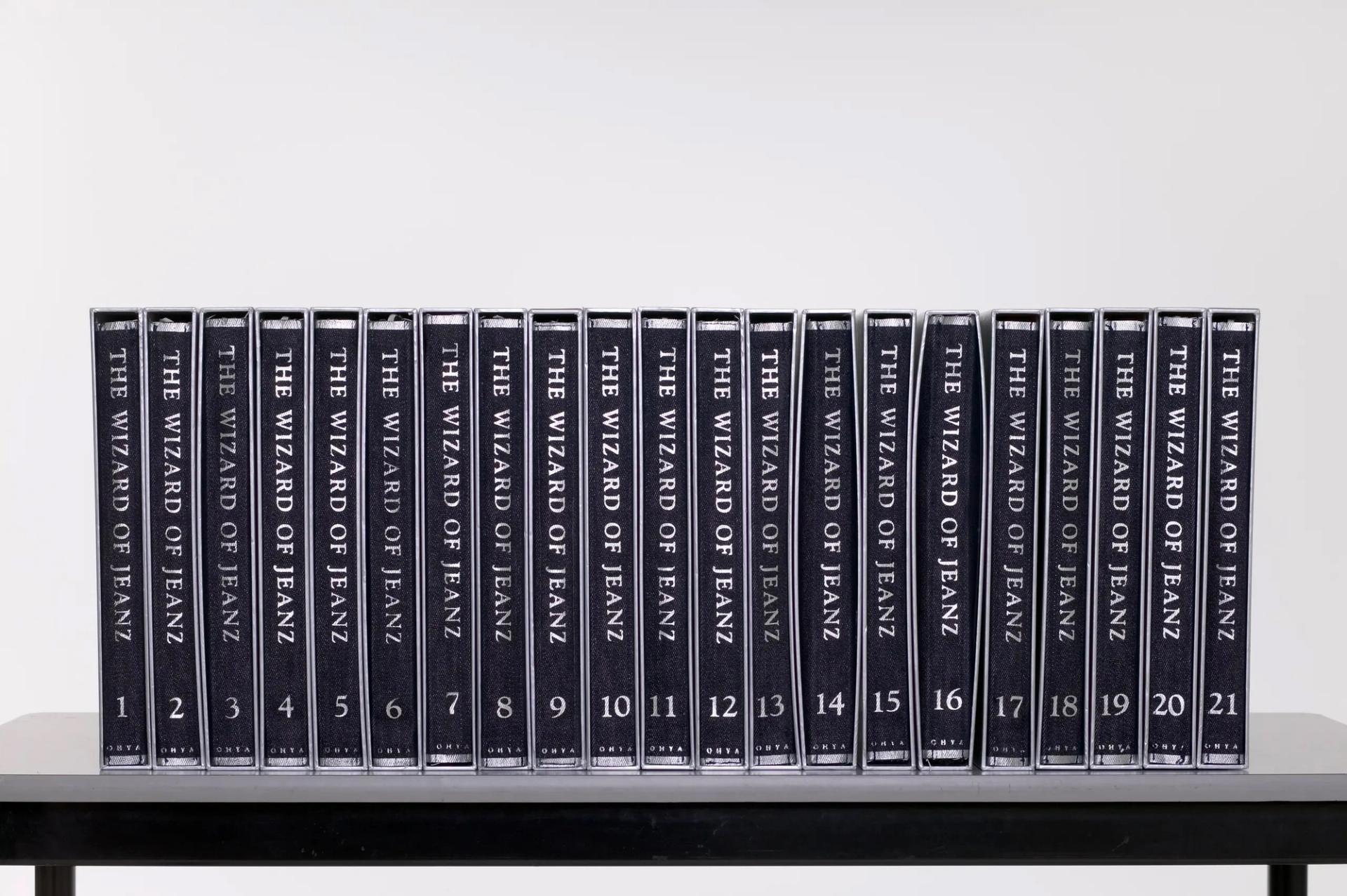 The 'Wizard of Jeanz' collection is a series of twenty-one fabric books that fold out to become different kinds of women's garments. The clothes are sewn into the inside 'spine' of a denim-look fabric booklet which can be folded to fit inside a silver cardboard slip cover. The slipcovers can be placed together and shelved as books. The text 'The Wizard of Jeanz', the volume number of each book and the designer's surname Ohya is printed along the exterior spine of the folded material in silver. The text 'The Wizard of Jeanz' and the surname Ohya is also embossed along the spine of the slip cover. As the series of garments progresses, the clothes change gradually from denim-style clothing to floral cotton.