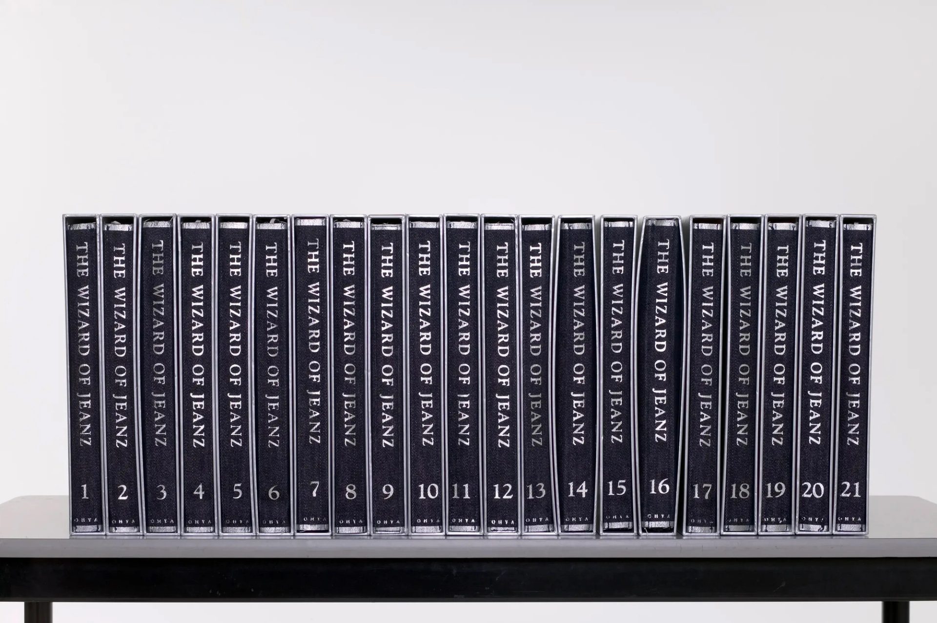 The 'Wizard of Jeanz' collection is a series of twenty-one fabric books that fold out to become different kinds of women's garments. The clothes are sewn into the inside 'spine' of a denim-look fabric booklet which can be folded to fit inside a silver cardboard slip cover. The slipcovers can be placed together and shelved as books. The text 'The Wizard of Jeanz', the volume number of each book and the designer's surname Ohya is printed along the exterior spine of the folded material in silver. The text 'The Wizard of Jeanz' and the surname Ohya is also embossed along the spine of the slip cover. As the series of garments progresses, the clothes change gradually from denim-style clothing to floral cotton.
