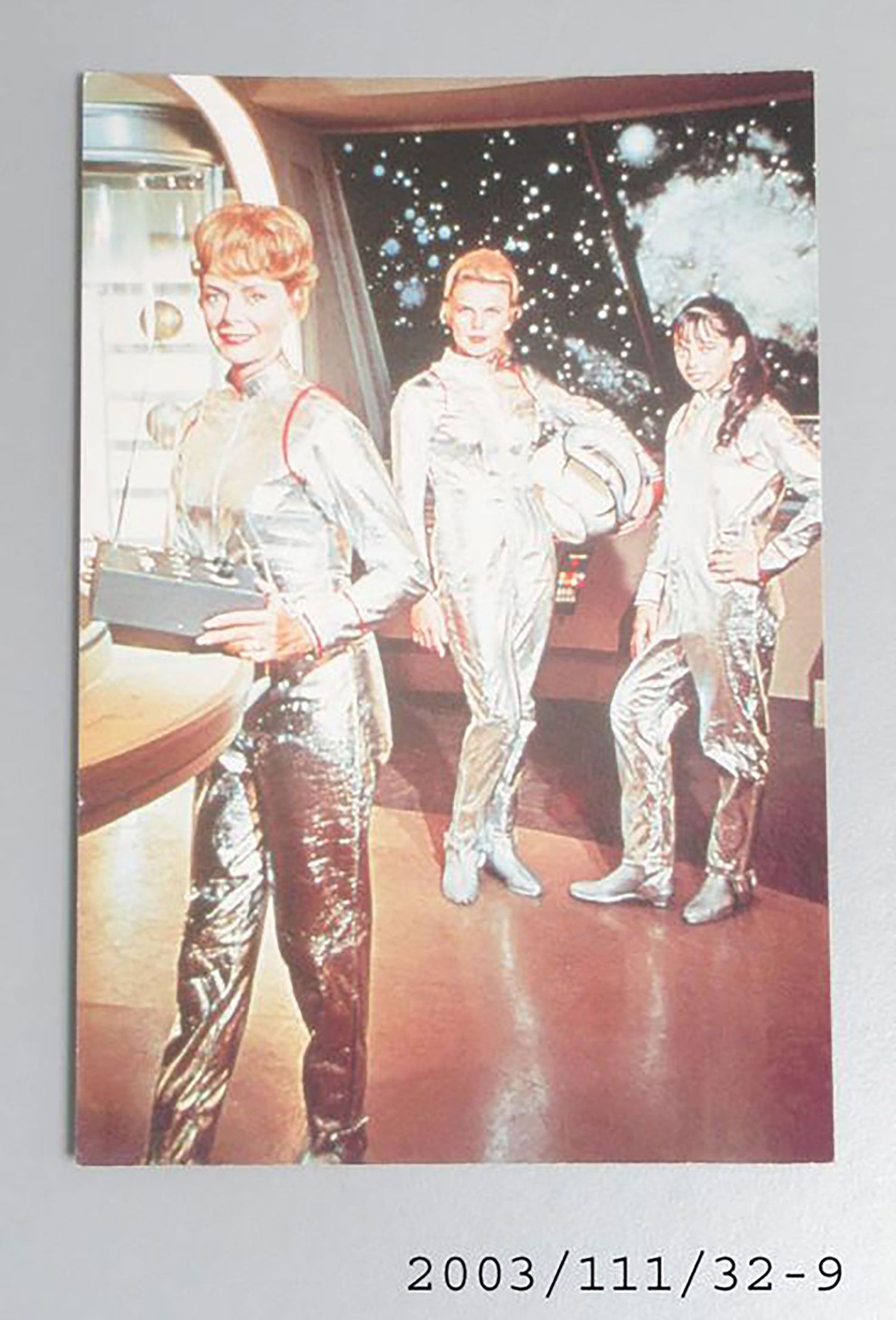Postcard, colour photographic print, licensed from the television show 'Lost in Space', cardboard, made by MA, Australia, 1980-1989. The postcard is made of cardboard and is printed in a portrait format. The postcard has a colour photographic print of 'Lost in Space' characters Maureen, Judy and Penny Robinson. They are standing inside a space ship and are wearing silver spacesuits.
