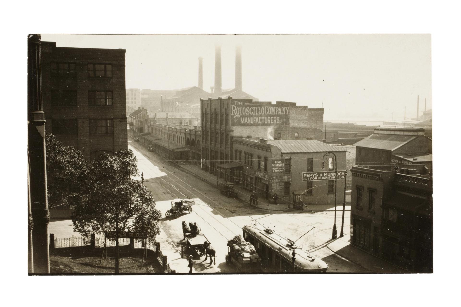 Old black-and-white photograph of a city street, with a tram and horse-drawn carriage in the foreground, and three tall industrial chimneys in the background.