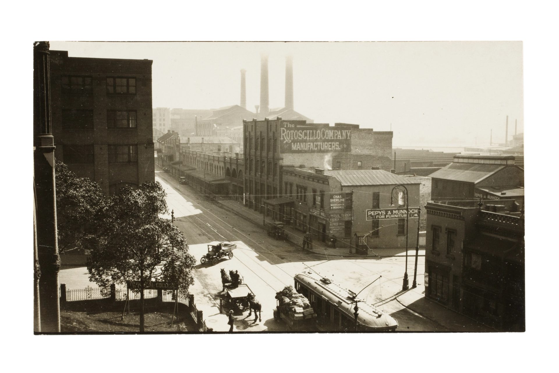 Old black-and-white photograph of a city street, with a tram and horse-drawn carriage in the foreground, and three tall industrial chimneys in the background.