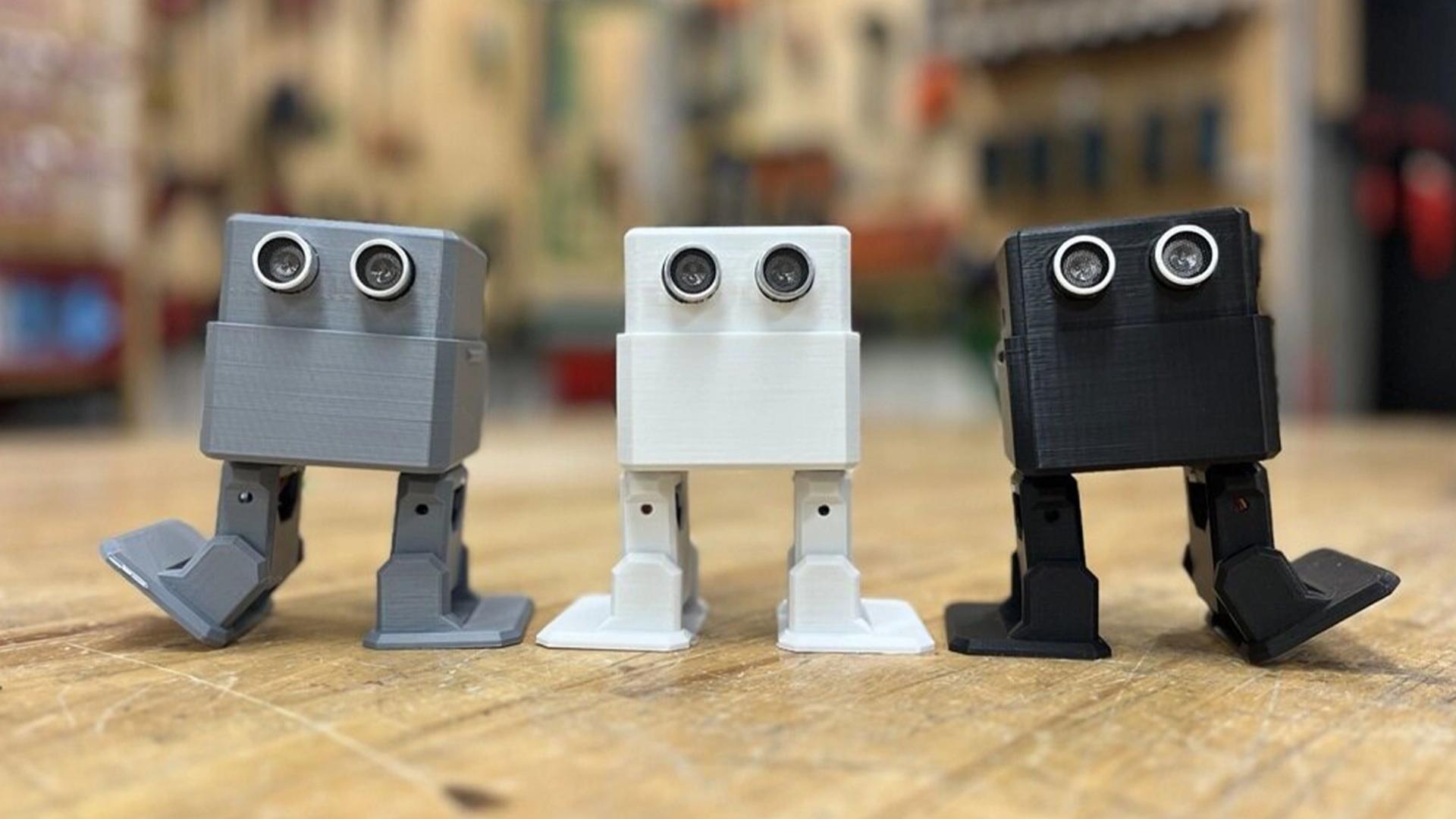 Three square robots – coloured (from left to right) grey, white and black, and with legs and eyes – stand on a wooden platform with a blurred background.