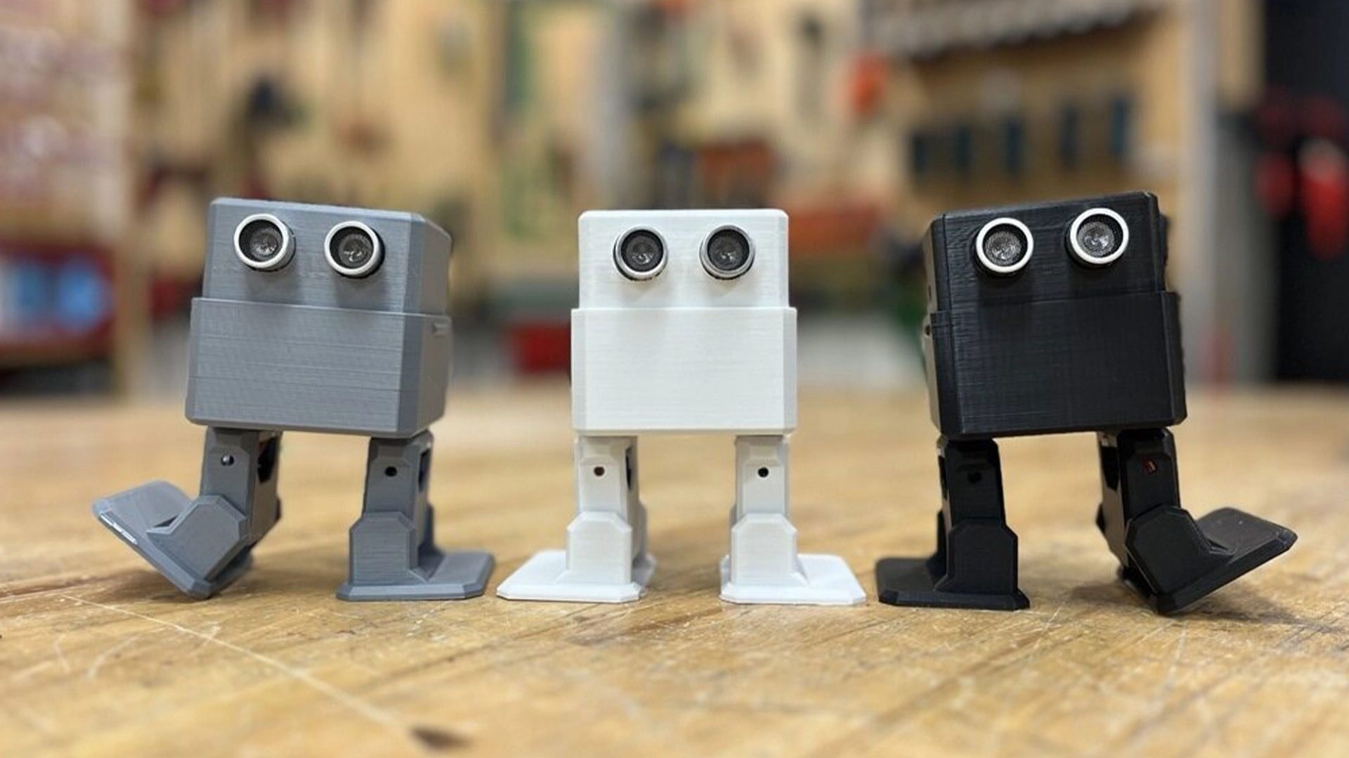 Three square robots – coloured (from left to right) grey, white and black, and with legs and eyes – stand on a wooden platform with a blurred background.