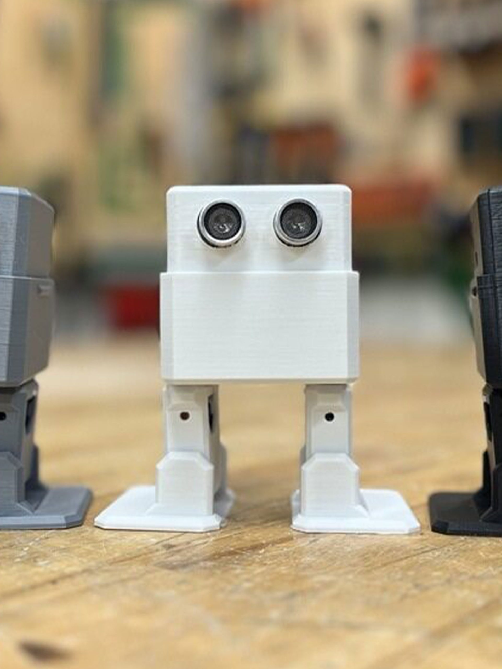 Three square robots – coloured (from left to right) grey, white and black, and with legs and eyes – stand on a wooden platform with a blurred background.