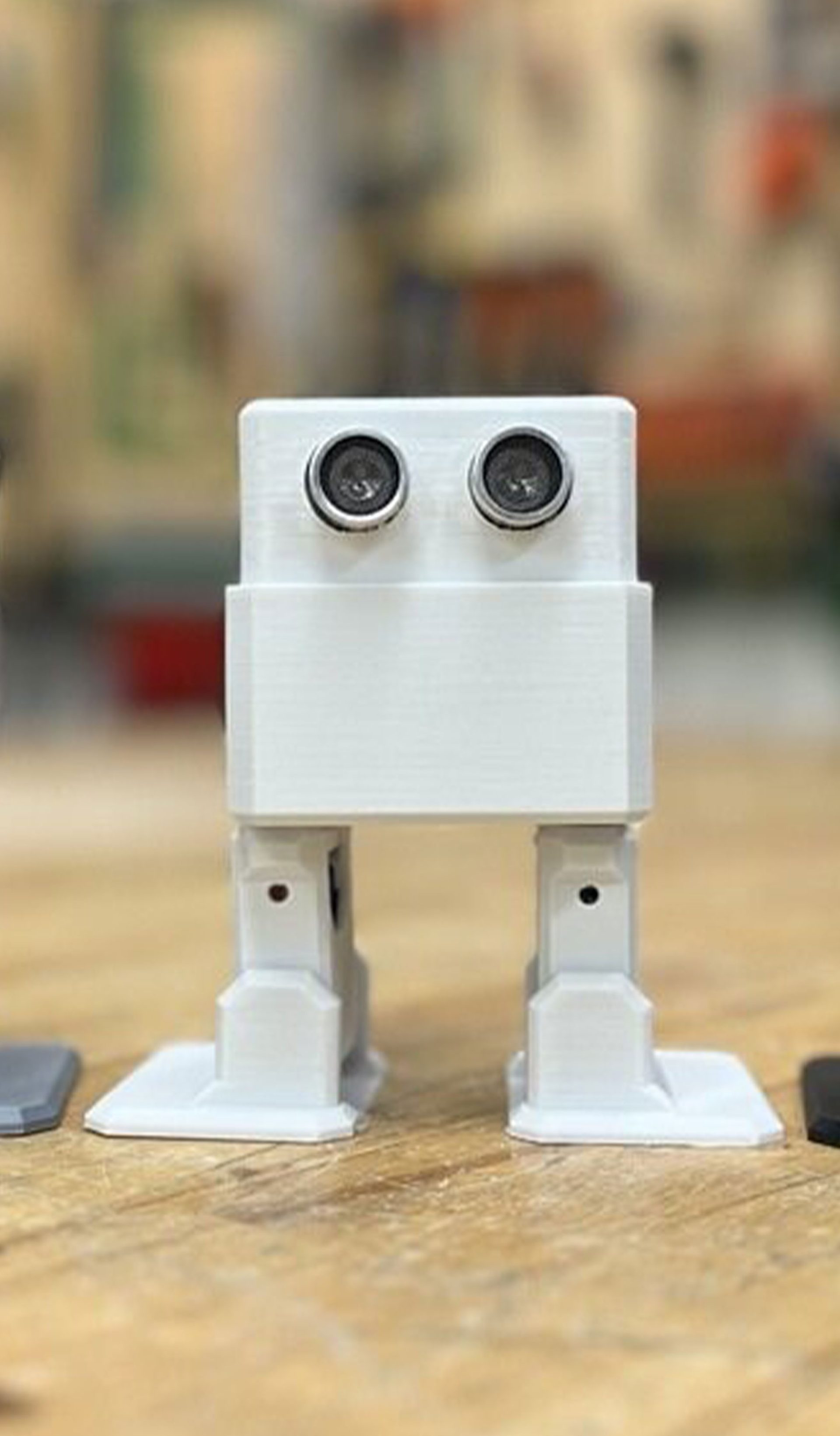 Three square robots – coloured (from left to right) grey, white and black, and with legs and eyes – stand on a wooden platform with a blurred background.