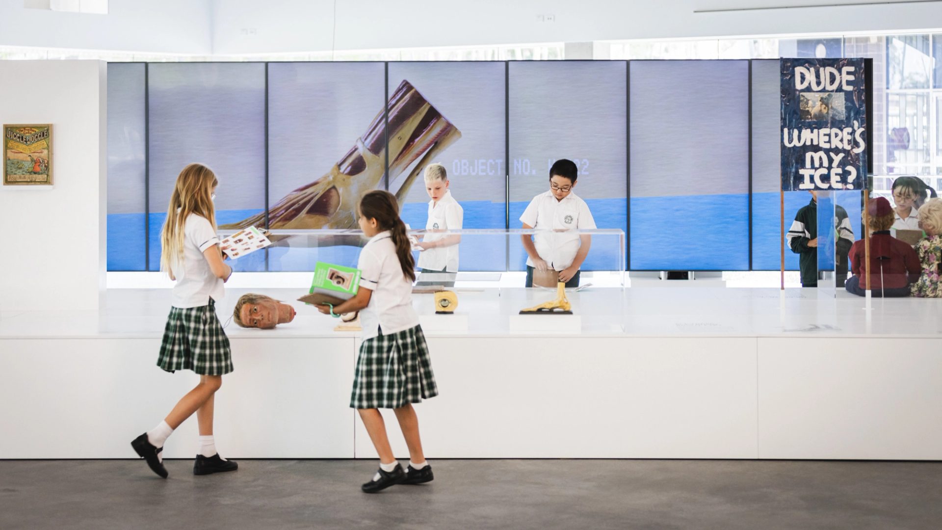 School children in uniforms explore a contemporary art installation in a bright gallery space. Several students in white shirts, plaid skirts or shorts, and black shoes examine quirky objects inside clear acrylic vitrines, including a severed head-like sculpture, a yellow oozing form, and other surreal items on white pedestals. Behind them, large floor-to-ceiling LED screens display close-up images of a giant metallic or organic object against a blue oceanic background, with text labels such as "OBJECT NO. 002". One child holds a green booklet, another peers into a display. A tall vertical sign reads "DUDE WHERE'S MY ICE?" in bold white letters on a dark background with a small photo. A vintage-style framed poster hangs on the left wall.
