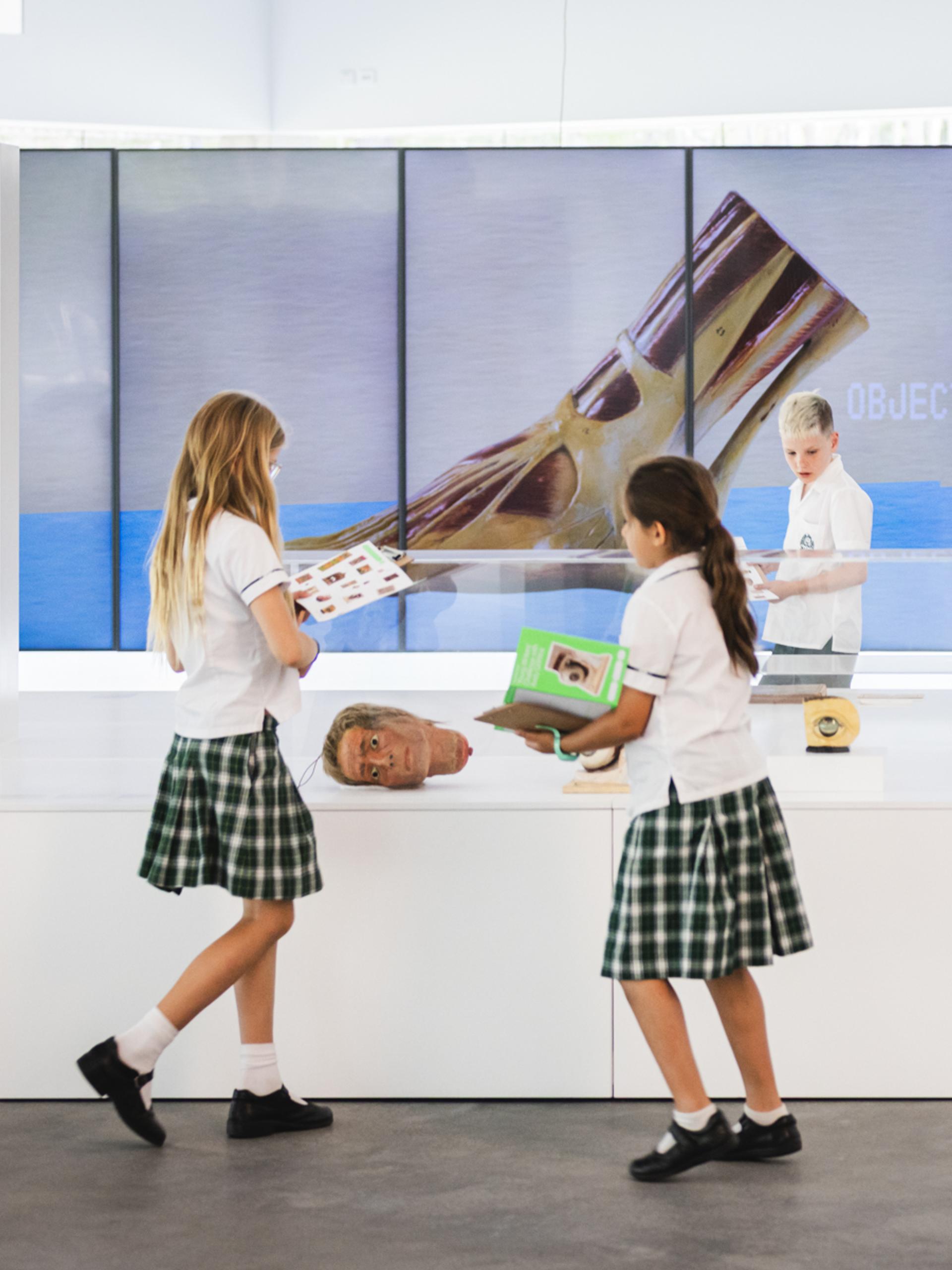 School children in uniforms explore a contemporary art installation in a bright gallery space. Several students in white shirts, plaid skirts or shorts, and black shoes examine quirky objects inside clear acrylic vitrines, including a severed head-like sculpture, a yellow oozing form, and other surreal items on white pedestals. Behind them, large floor-to-ceiling LED screens display close-up images of a giant metallic or organic object against a blue oceanic background, with text labels such as "OBJECT NO. 002". One child holds a green booklet, another peers into a display. A tall vertical sign reads "DUDE WHERE'S MY ICE?" in bold white letters on a dark background with a small photo. A vintage-style framed poster hangs on the left wall.