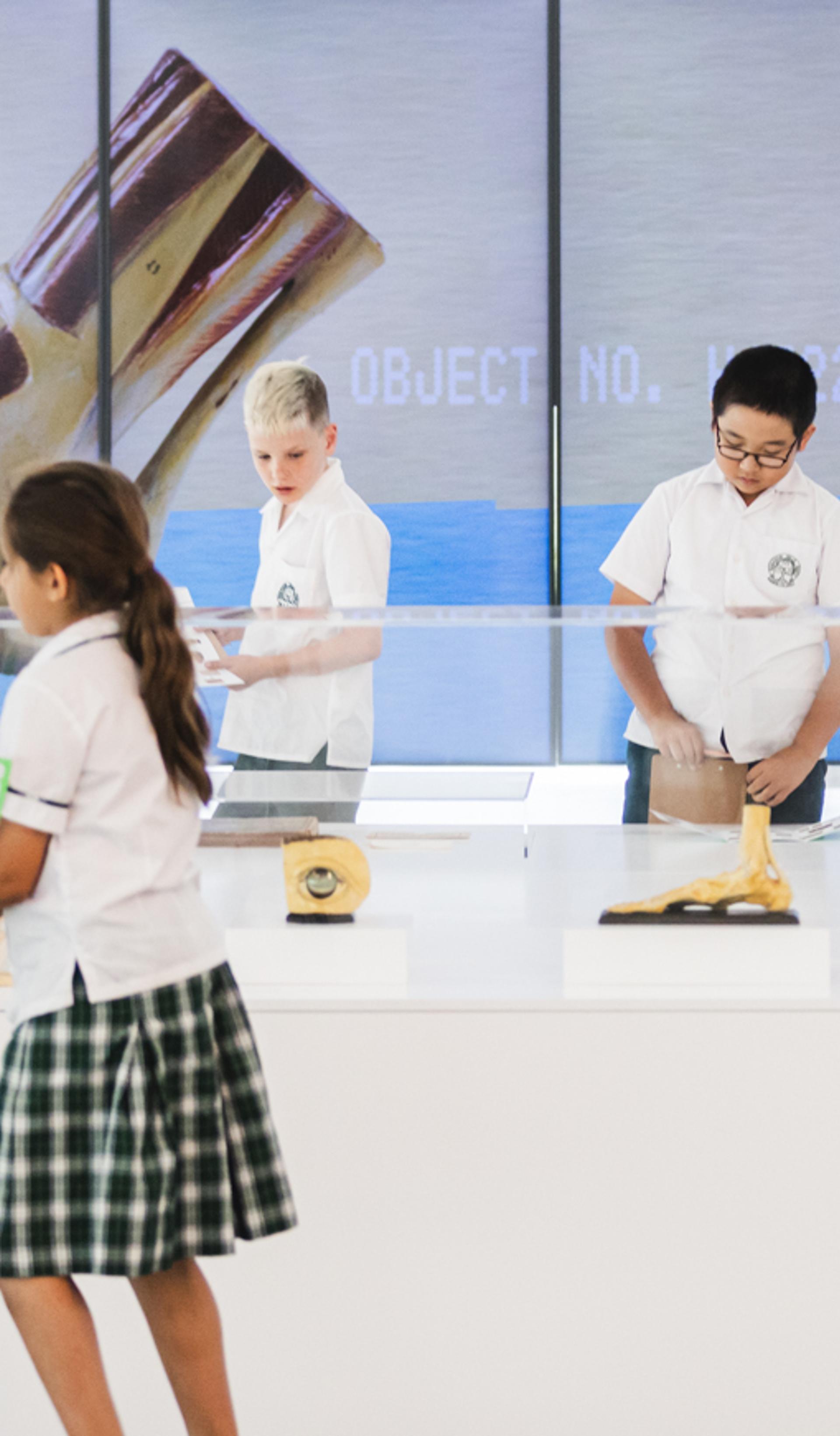 School children in uniforms explore a contemporary art installation in a bright gallery space. Several students in white shirts, plaid skirts or shorts, and black shoes examine quirky objects inside clear acrylic vitrines, including a severed head-like sculpture, a yellow oozing form, and other surreal items on white pedestals. Behind them, large floor-to-ceiling LED screens display close-up images of a giant metallic or organic object against a blue oceanic background, with text labels such as "OBJECT NO. 002". One child holds a green booklet, another peers into a display. A tall vertical sign reads "DUDE WHERE'S MY ICE?" in bold white letters on a dark background with a small photo. A vintage-style framed poster hangs on the left wall.