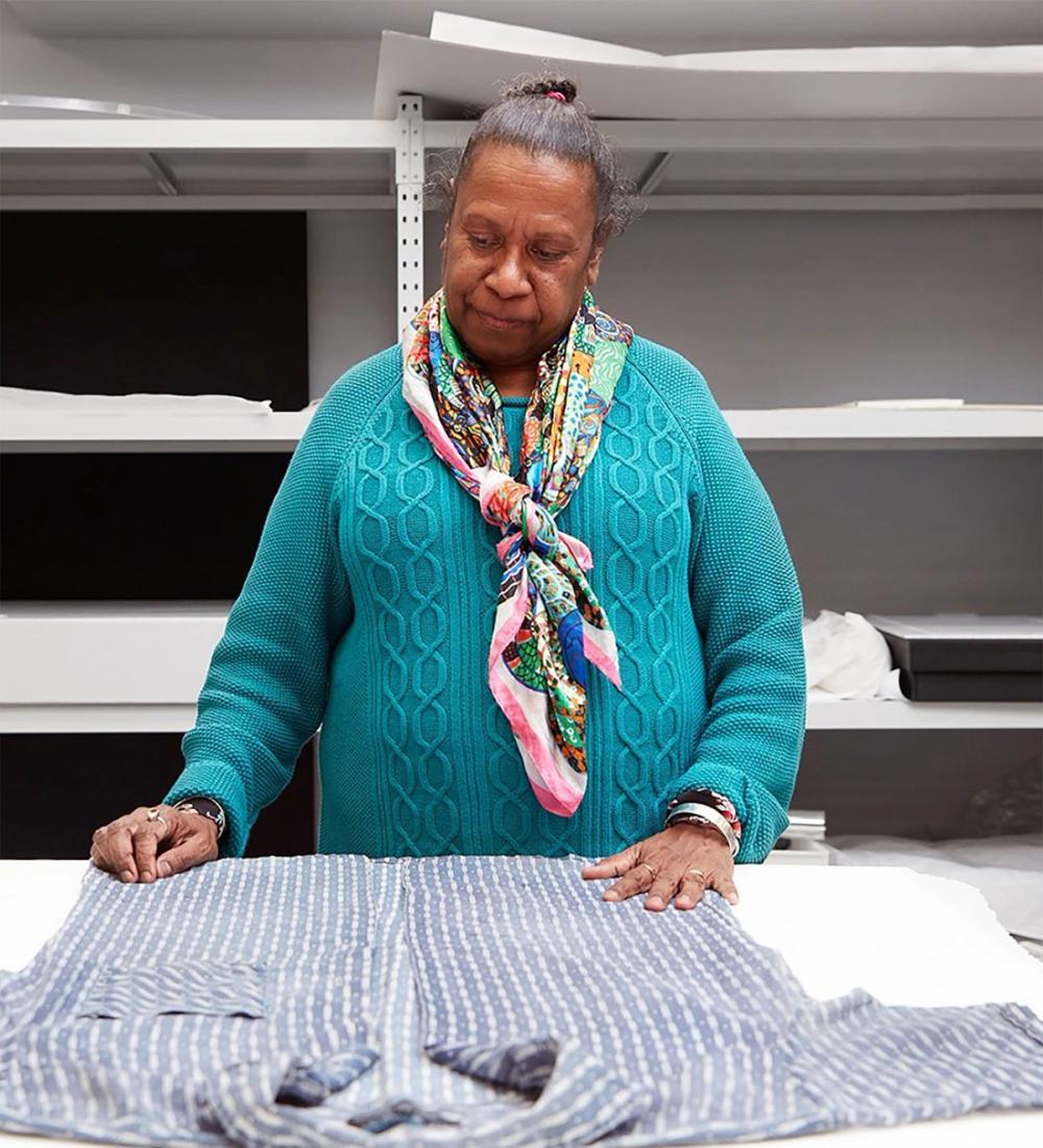 A middle aged Meriam woman looking at a flat lay shirt. The shirt is blue with a white vertical pattern on it. The woman is wearing a turquoise jumper.