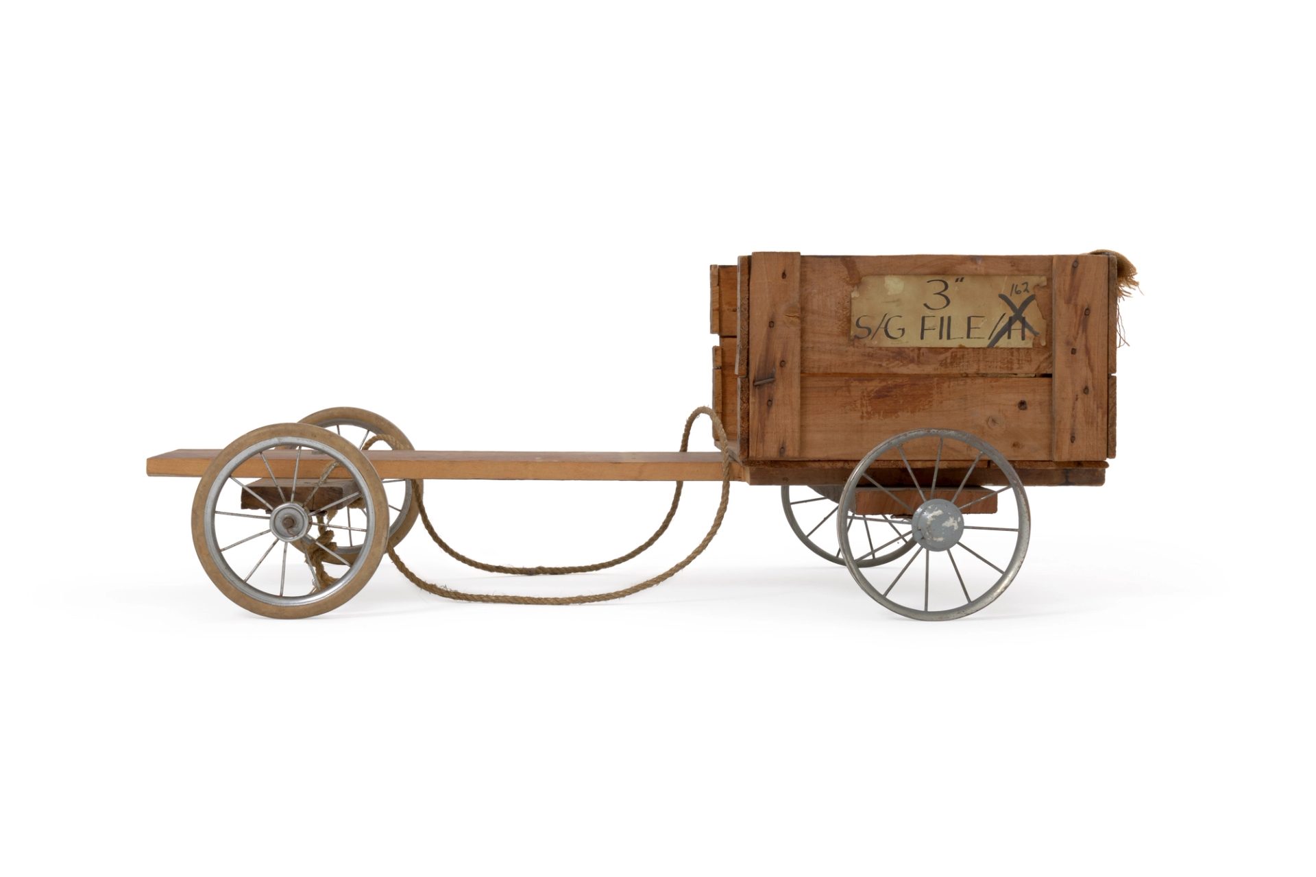 Billycart. The chassis of this billycart is made from wood. A plank forms the central axis. A wooden crate, cut down to four sides, forms the cabin and seat. A brown hessian bag has been draped over the back of this crate. Stuck on the left side of the crate is a piece of paper with the inscription; '3" 162 S/G FILE/H'.