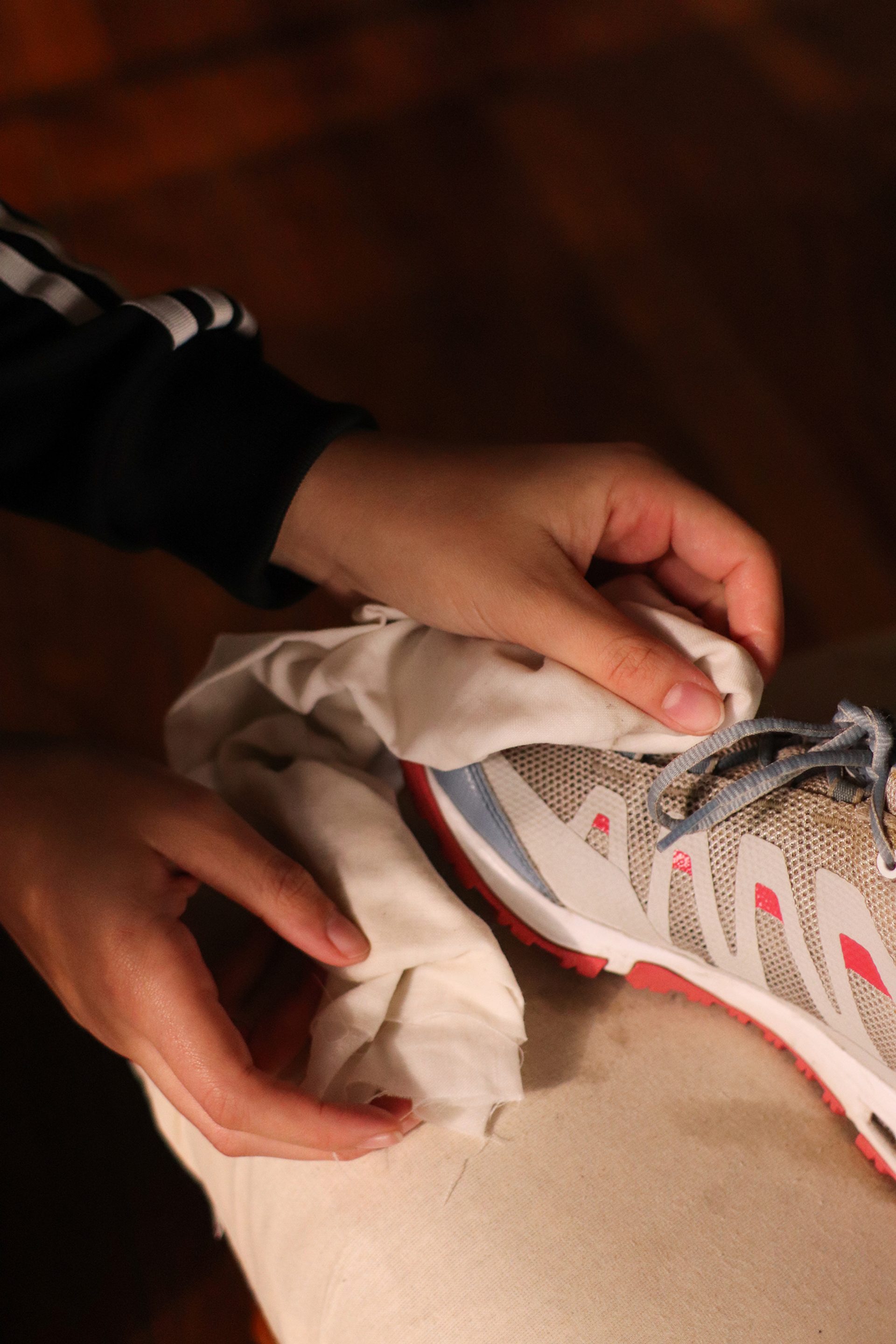 The hands of a young woman using a cloth to clean a sneaker on someone’s foot.