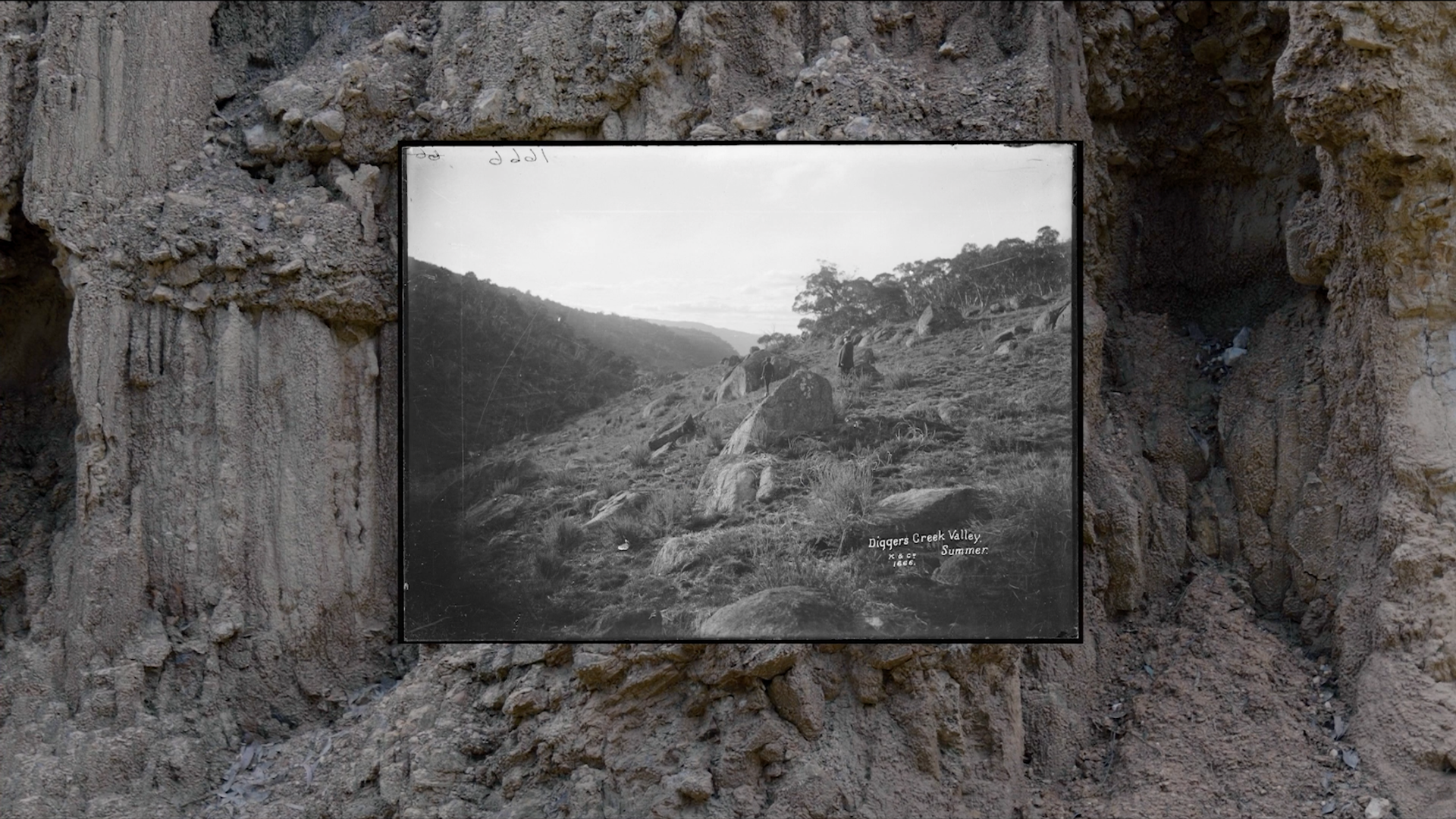 A black and white image of a large screen depicting a landscape. The screen is placed in front of a rock wall.