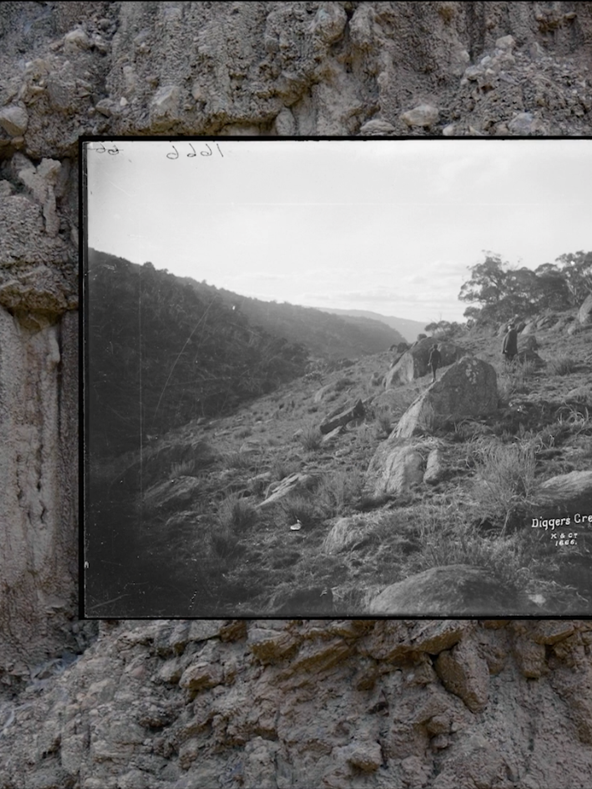 A black and white image of a large screen depicting a landscape. The screen is placed in front of a rock wall.