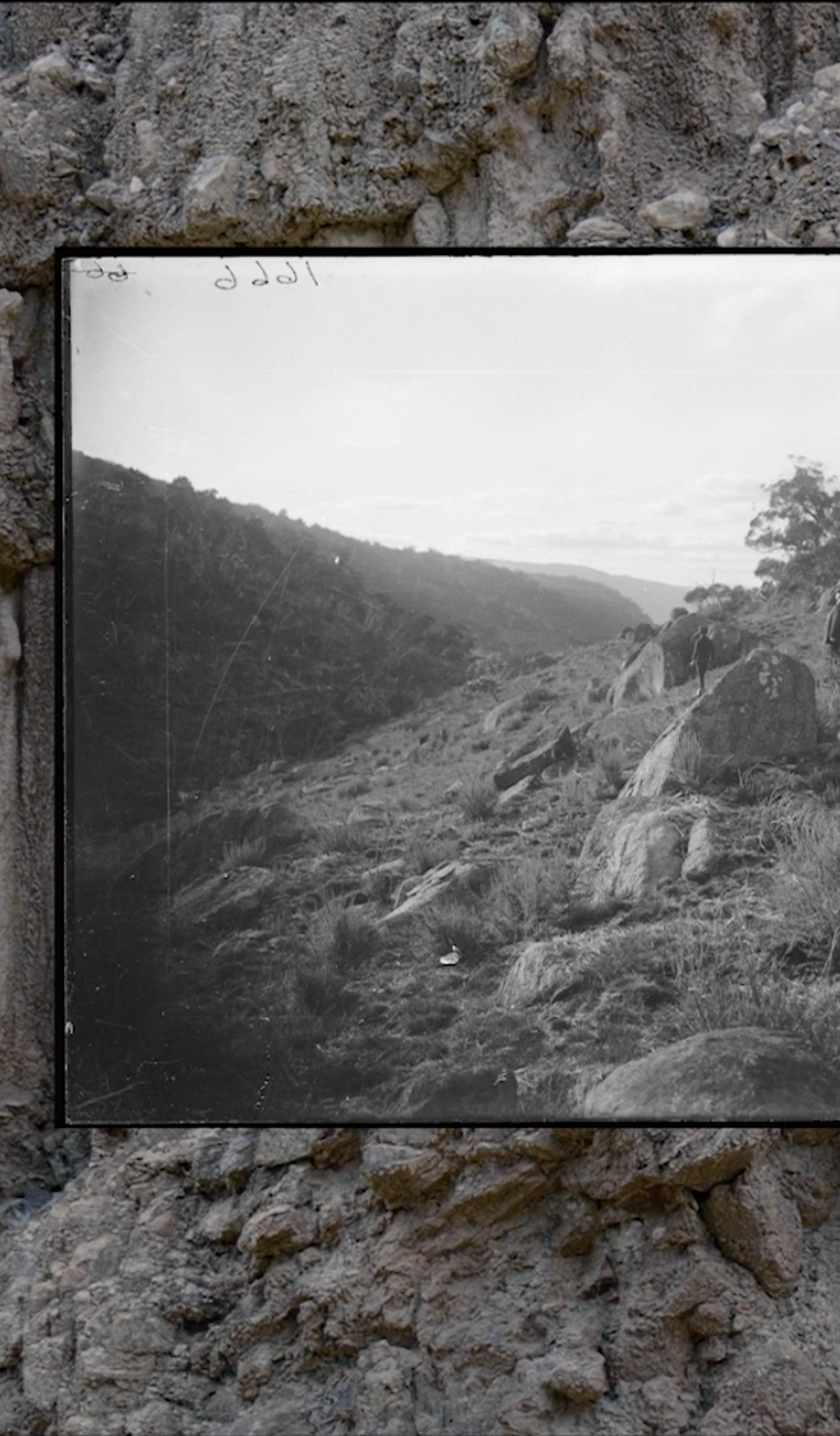 A black and white image of a large screen depicting a landscape. The screen is placed in front of a rock wall.