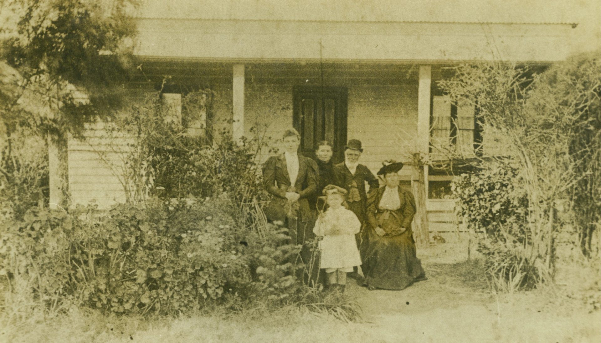A sepia family photograph from around the turn of the last century, picturing five figures, including an elderly couple in dark clothing and a child wearing white, standing at the garden entrance to a farmhouse.