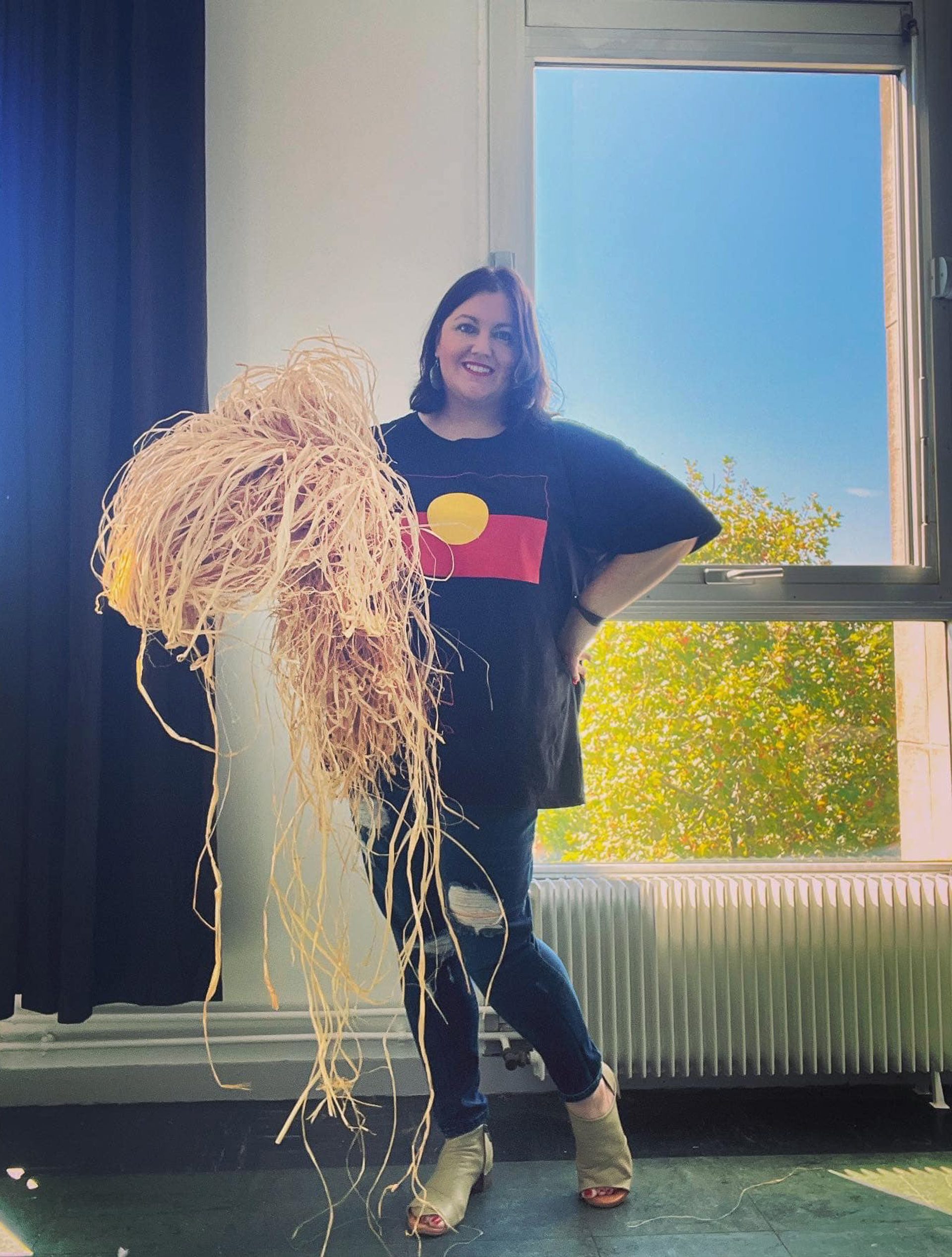 First nations woman stands holding raffia for weaving in front of window.