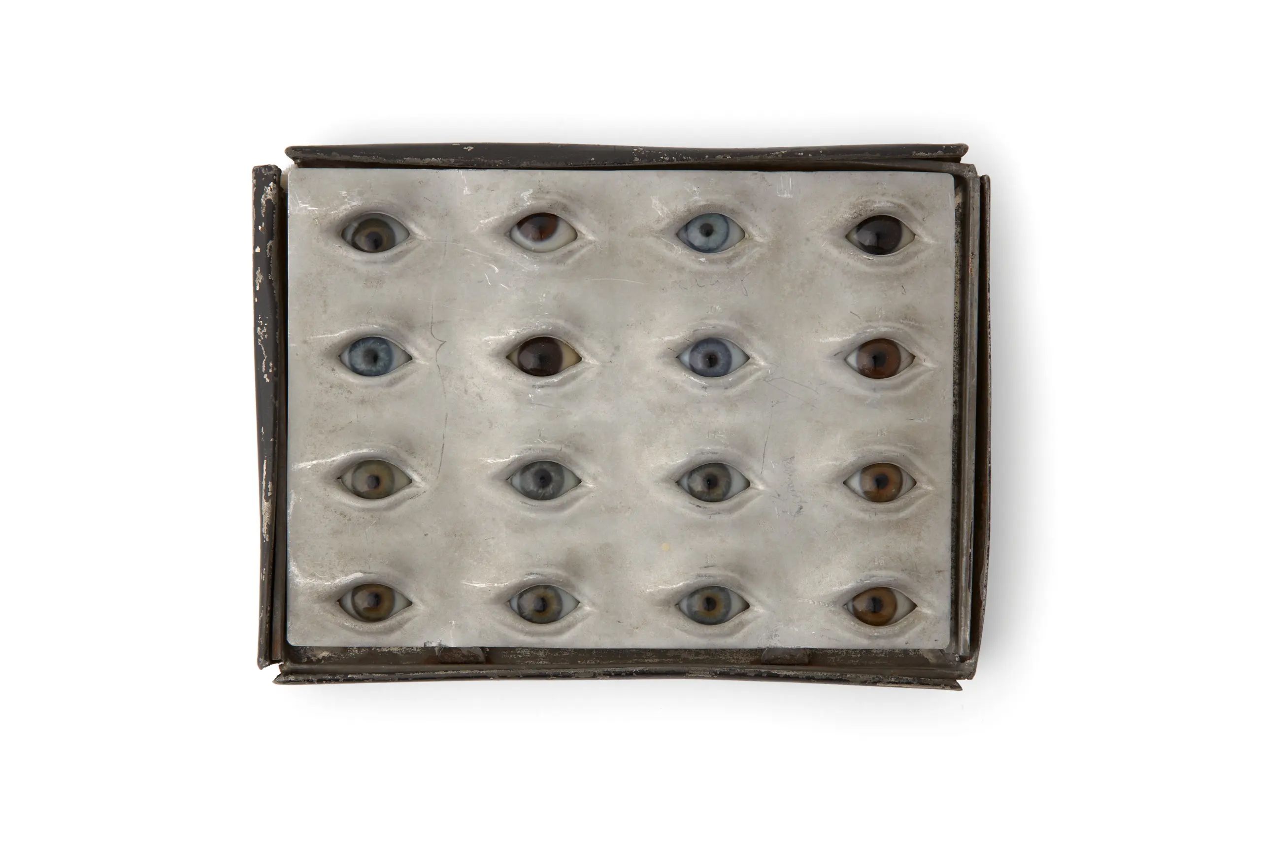 Photograph of board with spherical glass eyes with varying pupil sizes and colours including blue, hazel and brown. The glass eyes are housed within a tin box containing an insert with individual cut-outs for each eyeball.