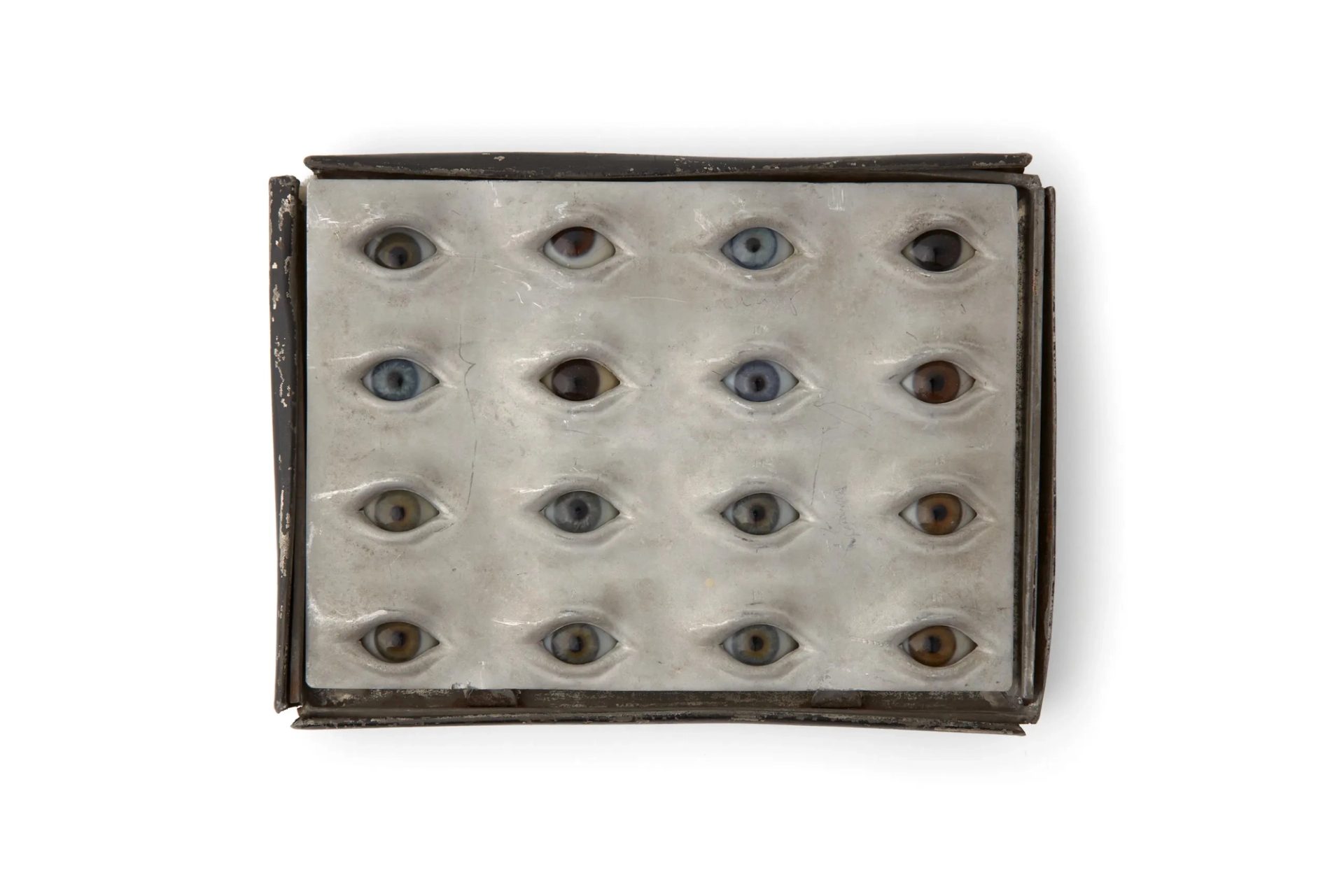 Photograph of board with spherical glass eyes with varying pupil sizes and colours including blue, hazel and brown. The glass eyes are housed within a tin box containing an insert with individual cut-outs for each eyeball.