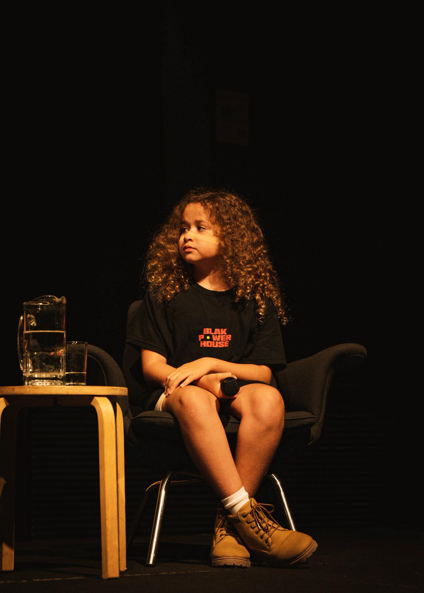 A girl is sitting on stage in a black chair. She is looking to her right and holding a microphone in her lap.