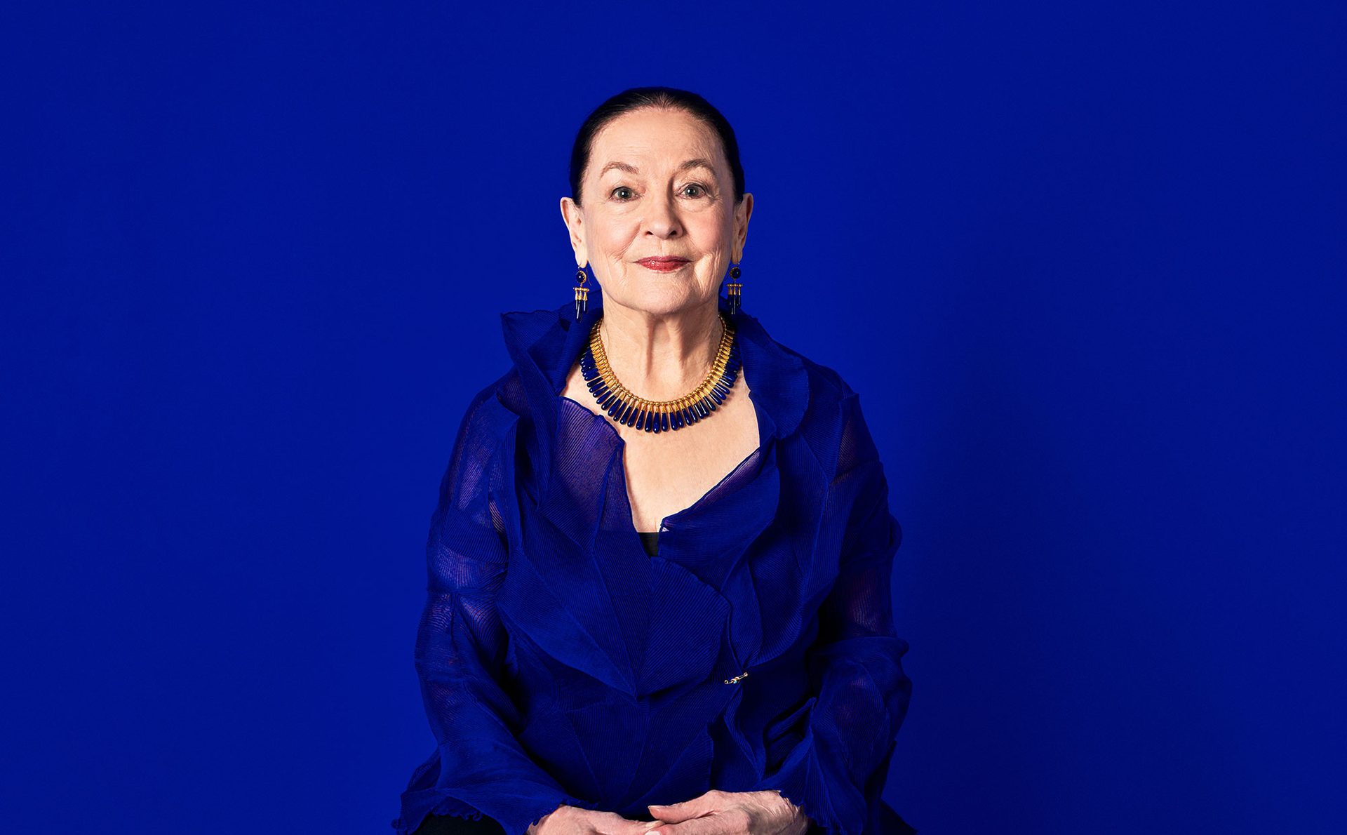 Portrait of a woman with dark hair slicked back wearing a blue and gold necklace with matching earrings.