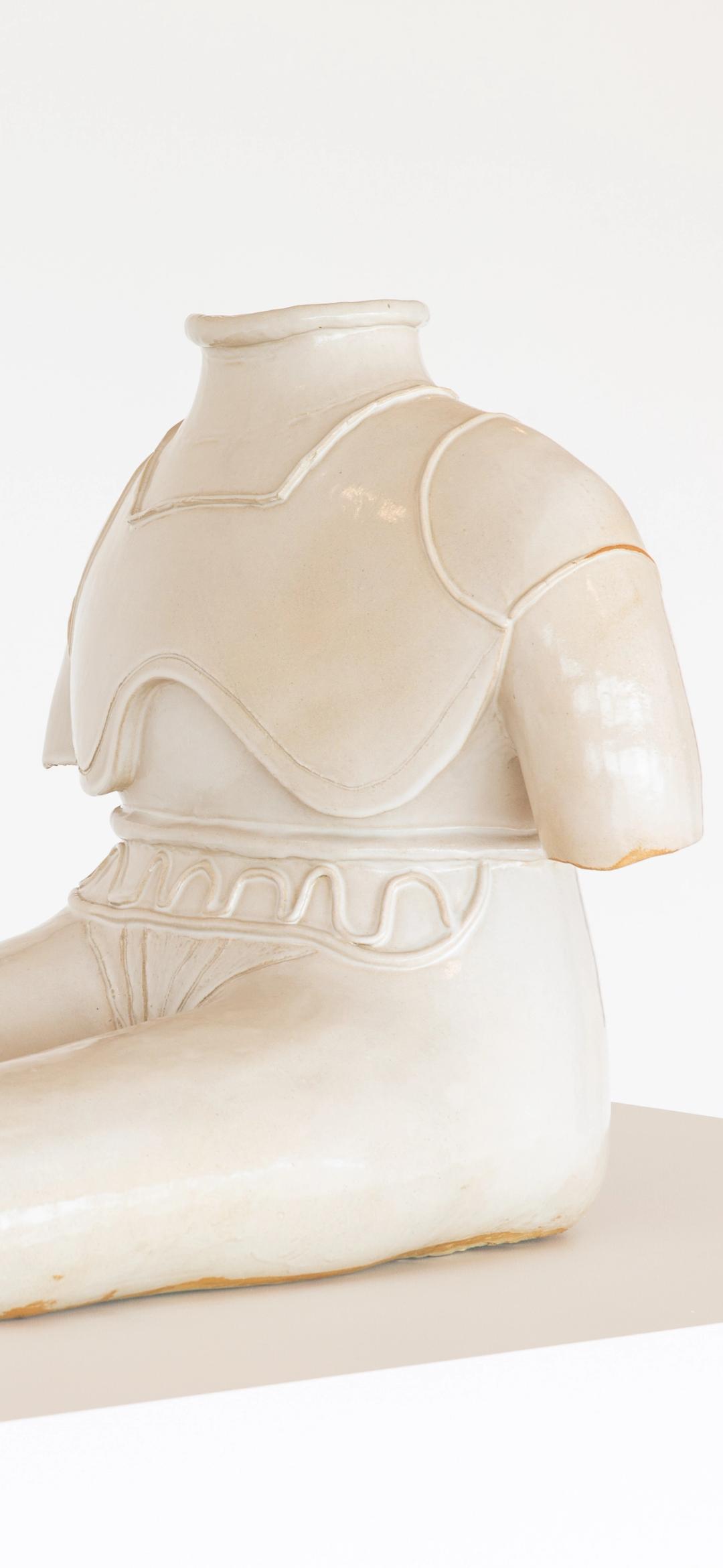 Statue of figure without feed or head sits with extended legs, glazed stoneware