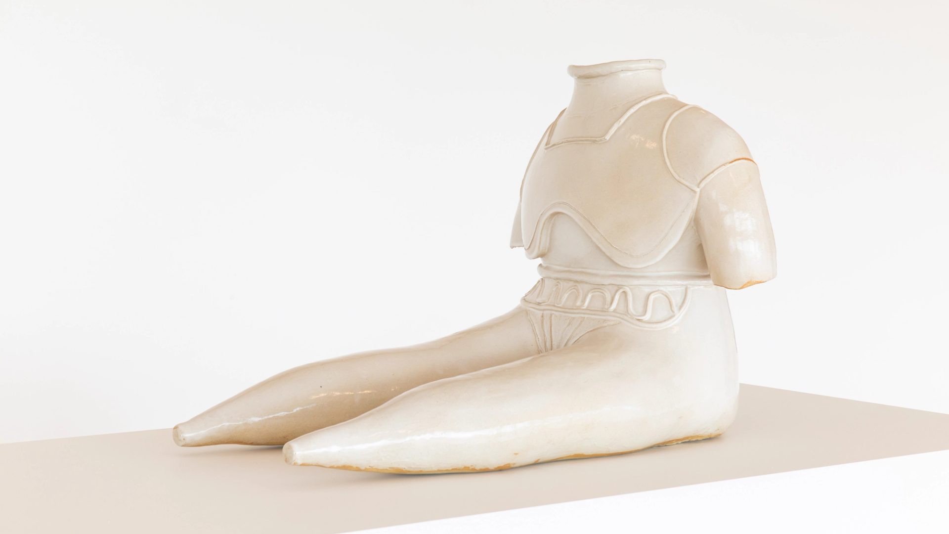 Statue of figure without feed or head sits with extended legs, glazed stoneware