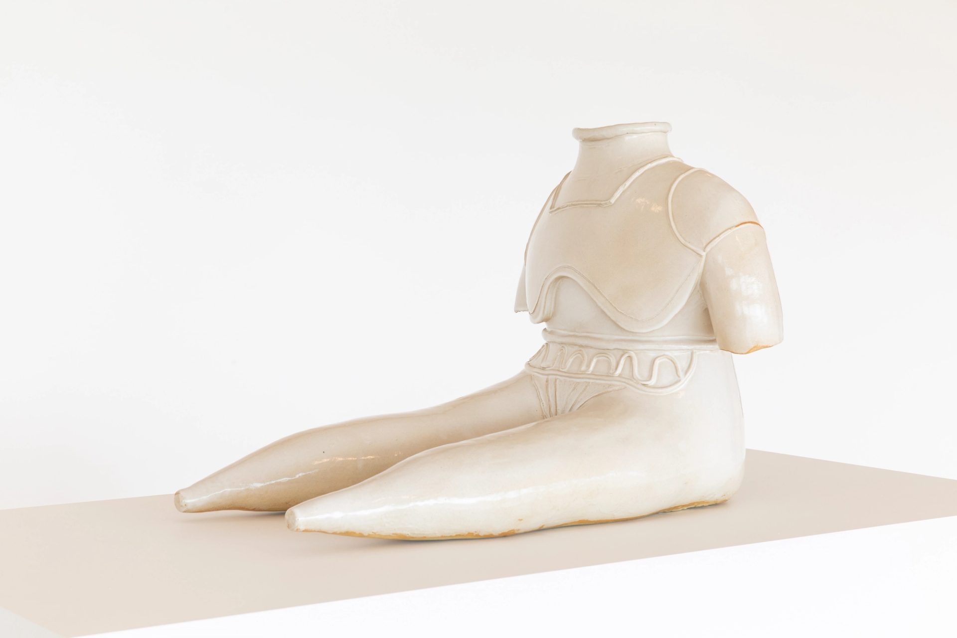 Statue of figure without feed or head sits with extended legs, glazed stoneware