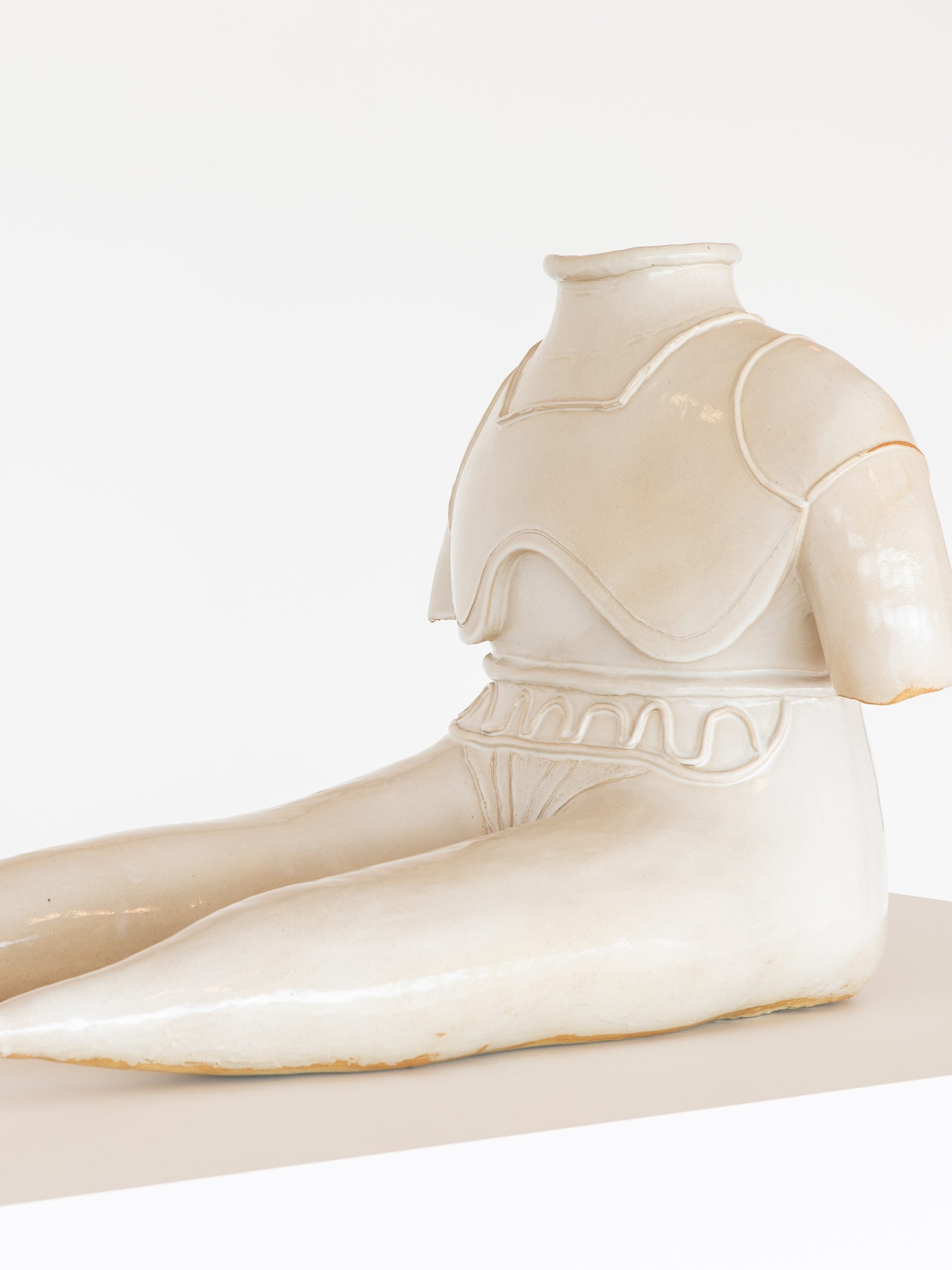 Statue of figure without feed or head sits with extended legs, glazed stoneware