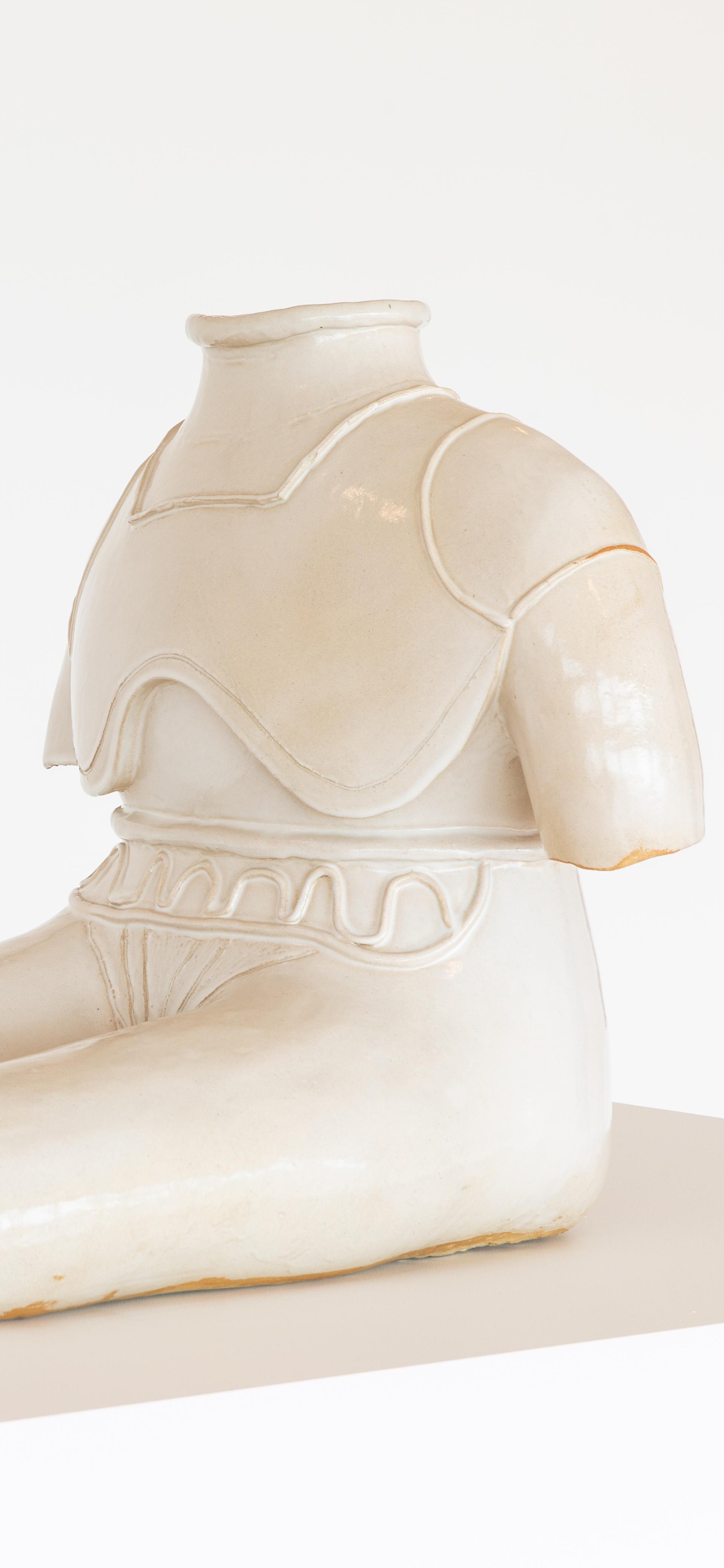 Statue of figure without feed or head sits with extended legs, glazed stoneware