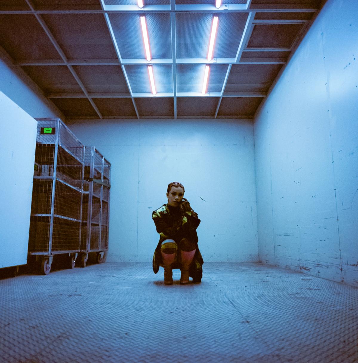 A young woman sits crouched in the middle of an industrial looking room.