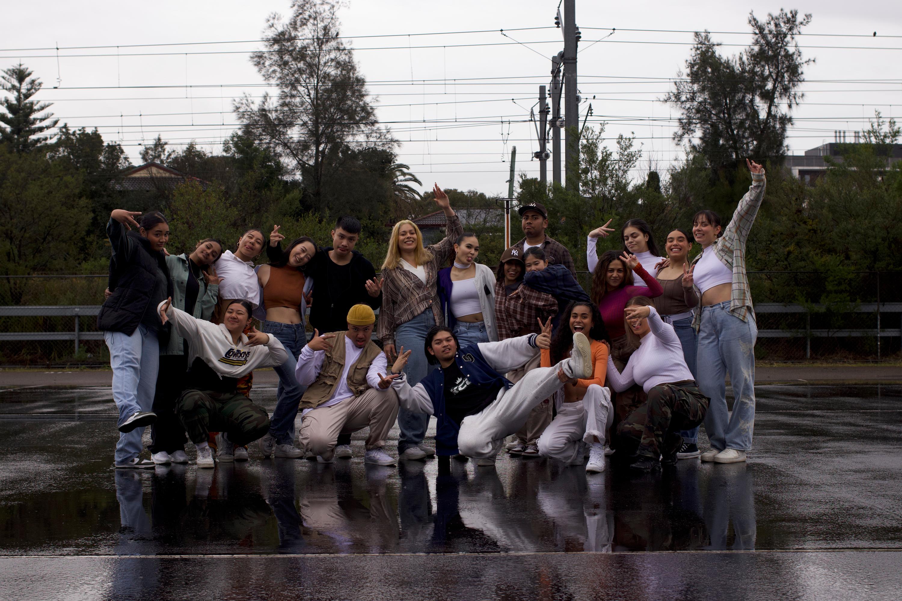 A group of young people dressed in street clothes pose in an empty carpark. The ground is wet. There are train lines visible in the background.