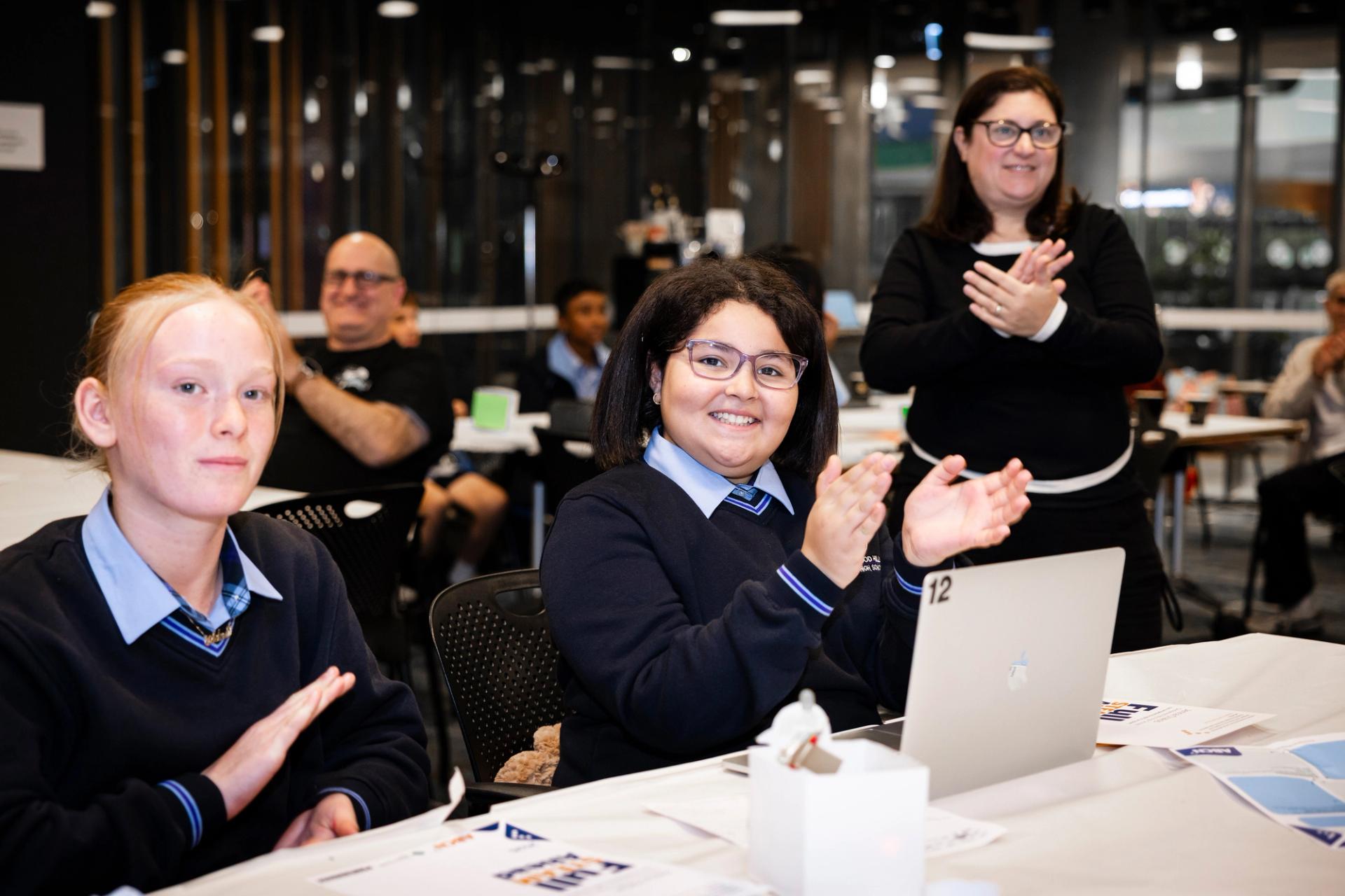 Photo of students sitting on a table applauding, there is a laptop in front of 1 student and a teacher standing in the background also applauding.