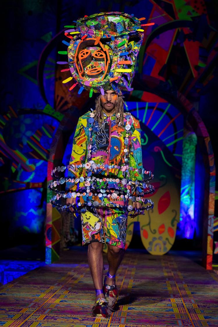 Runway model wearing colourful shorts and matching zip up jacket. four rings of buttons sit around their waist. A matching large headpiece with an orange face is on their head. Orange faces are painted on the model's cheeks.