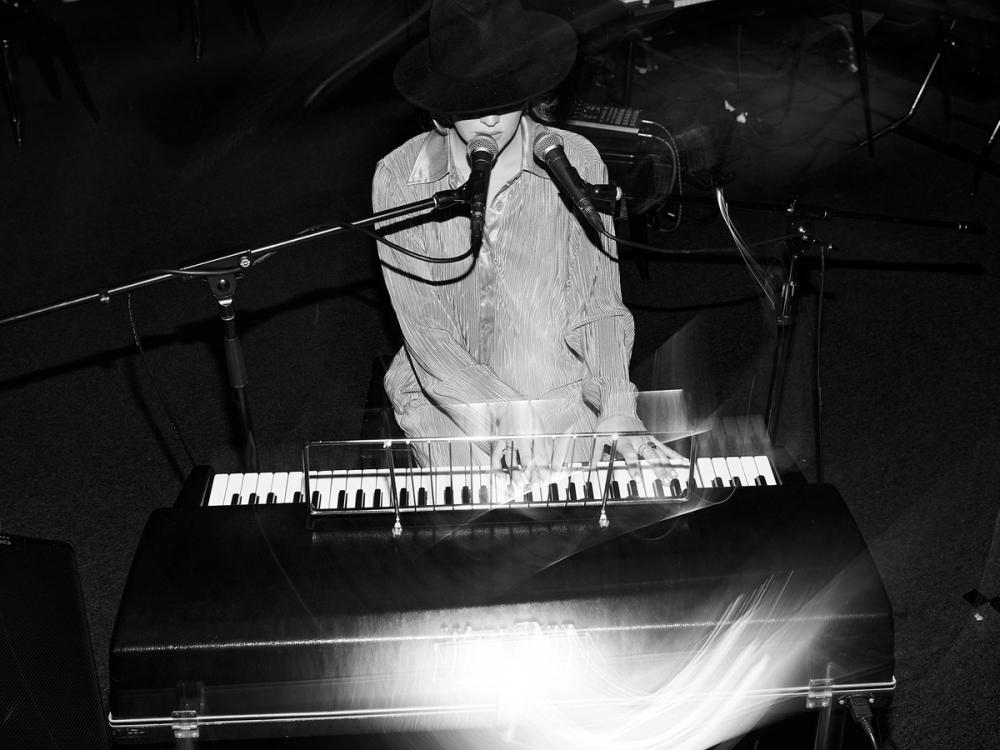 Performer sits at a keyboard singing into a microphone wearing a large hat that covers the eyes.