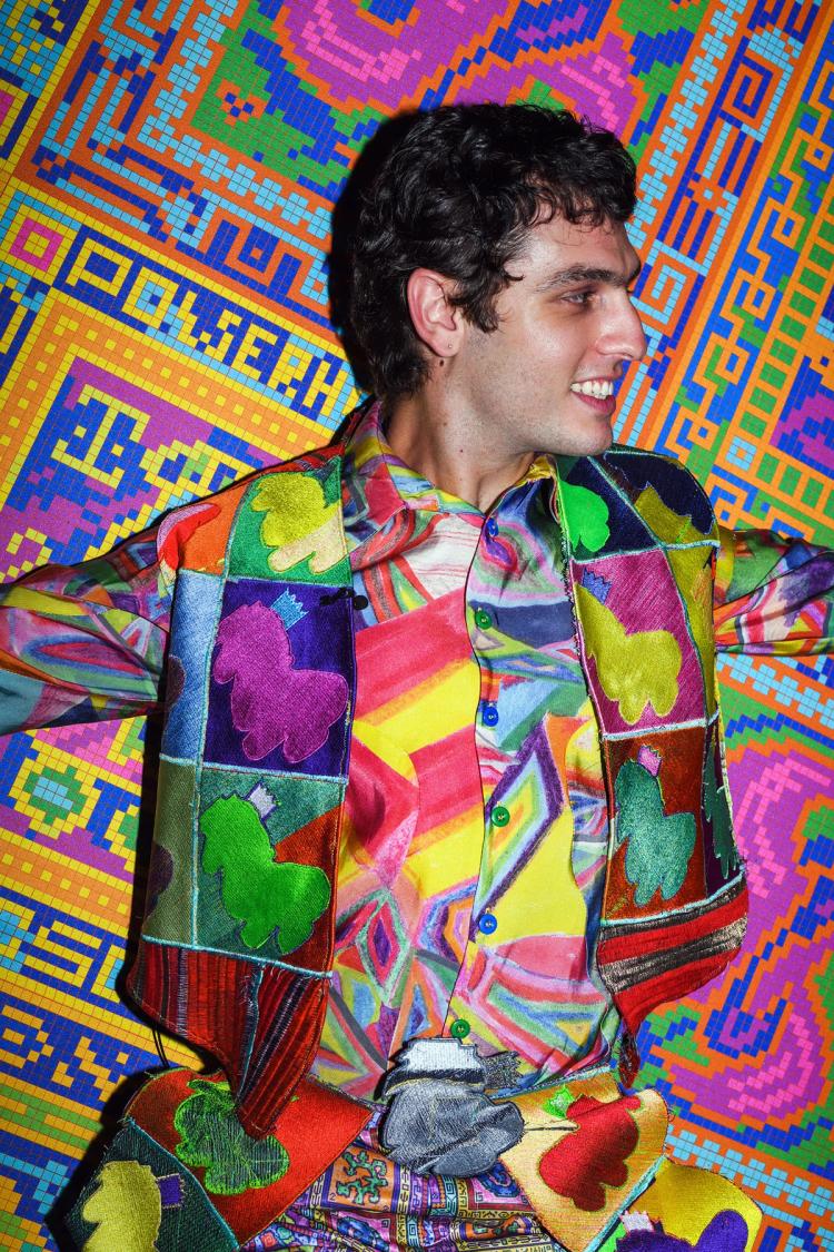 Jordan Gogos wearing a multicoloured shirt and jacket smiles looking to the right against a patterned background