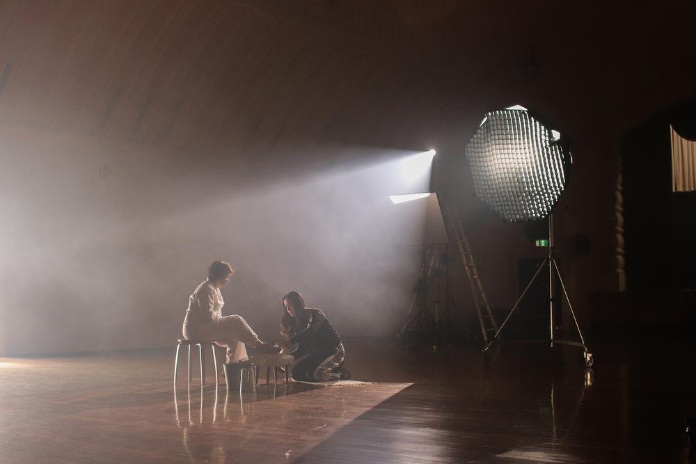 A film set in a spacious room with wooden floors. A young woman is on her knees cleaning an older woman’s shoe. The older woman sits on a stool. There is a film light visible.