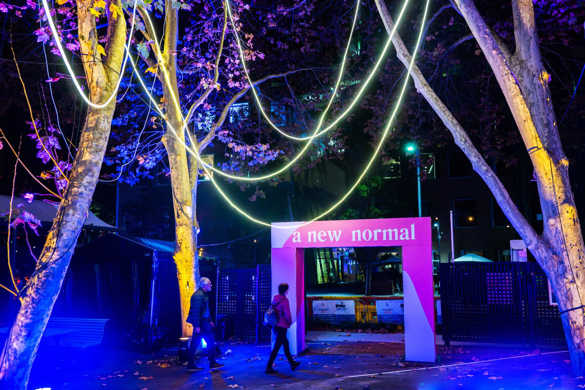 'A New Normal' entrance banner amongst colourfully lit trees in a courtyard.