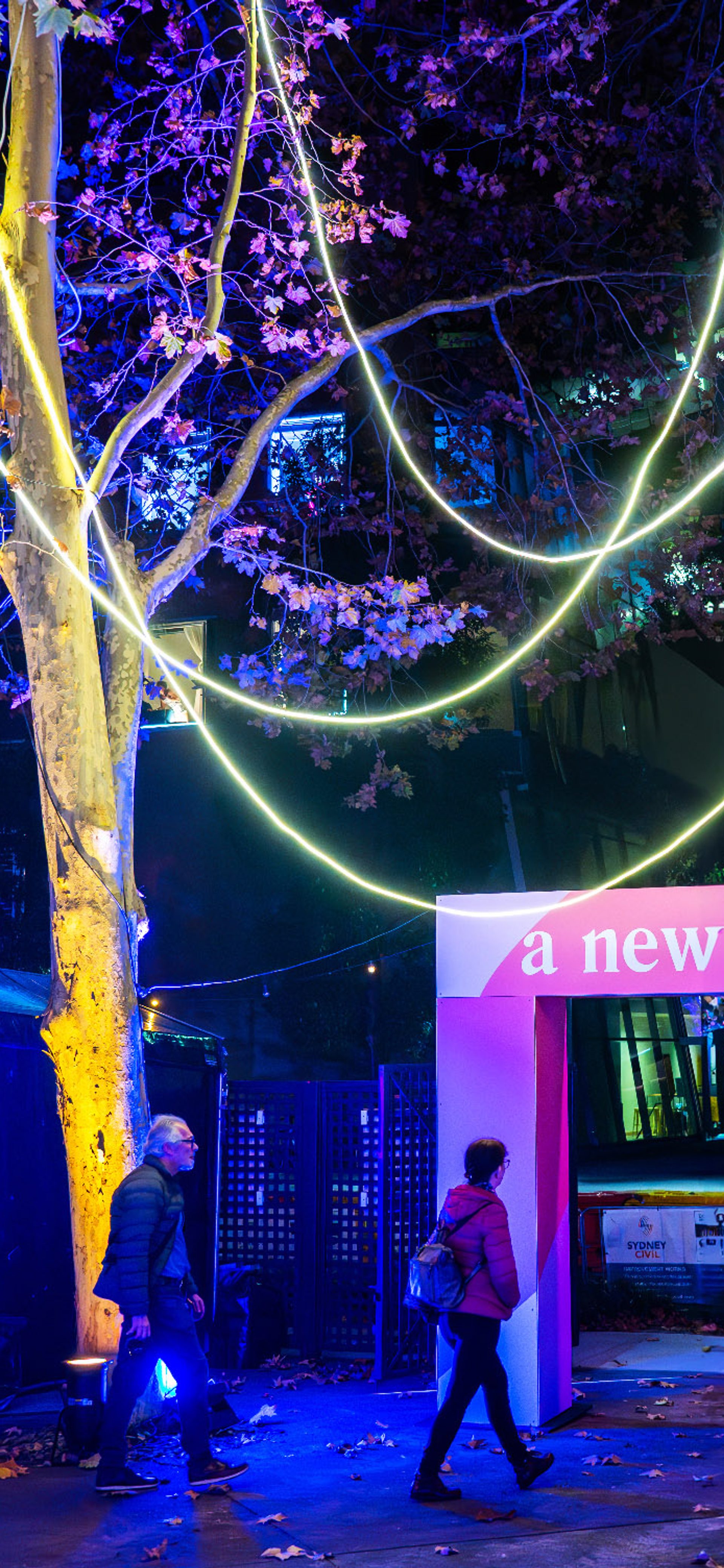 'A New Normal' entrance banner amongst colourfully lit trees in a courtyard.