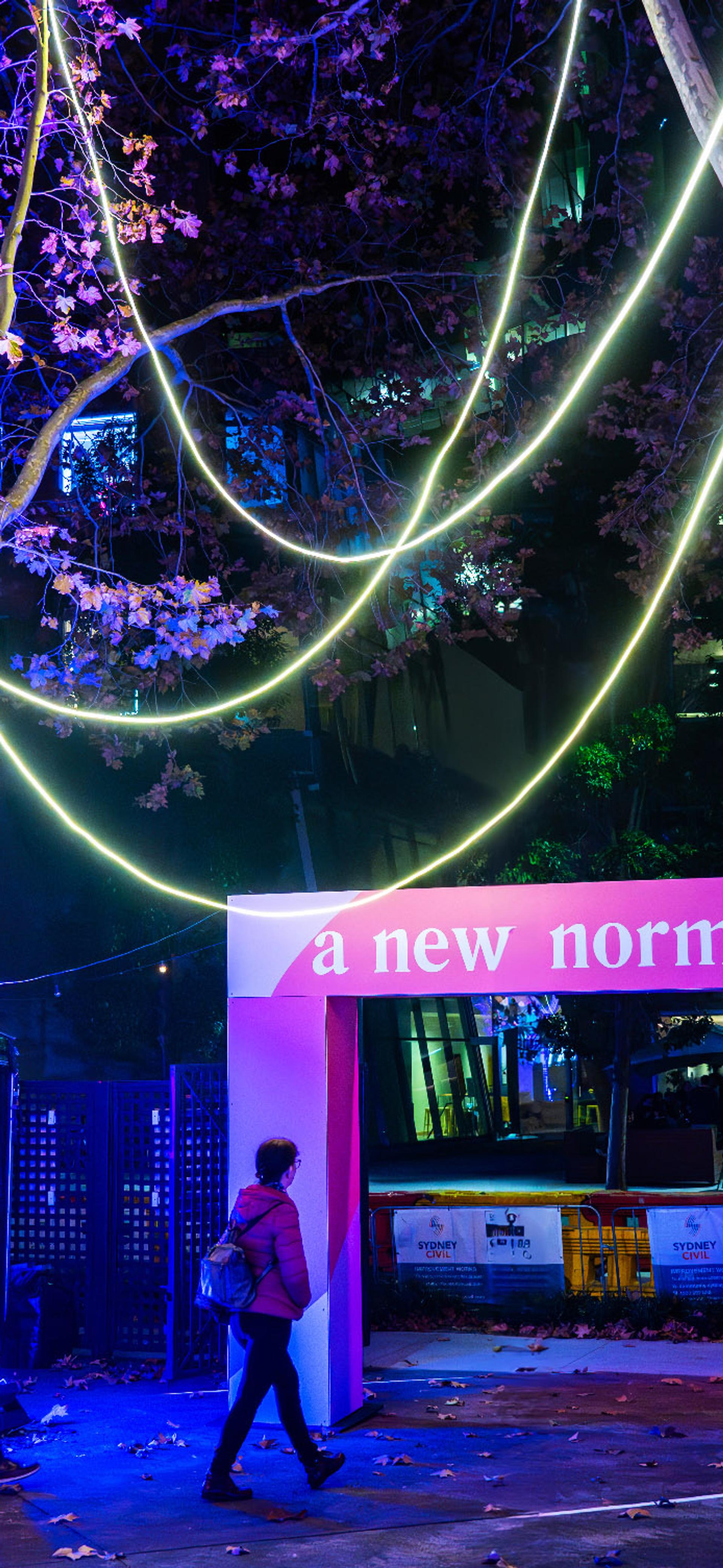 'A New Normal' entrance banner amongst colourfully lit trees in a courtyard.