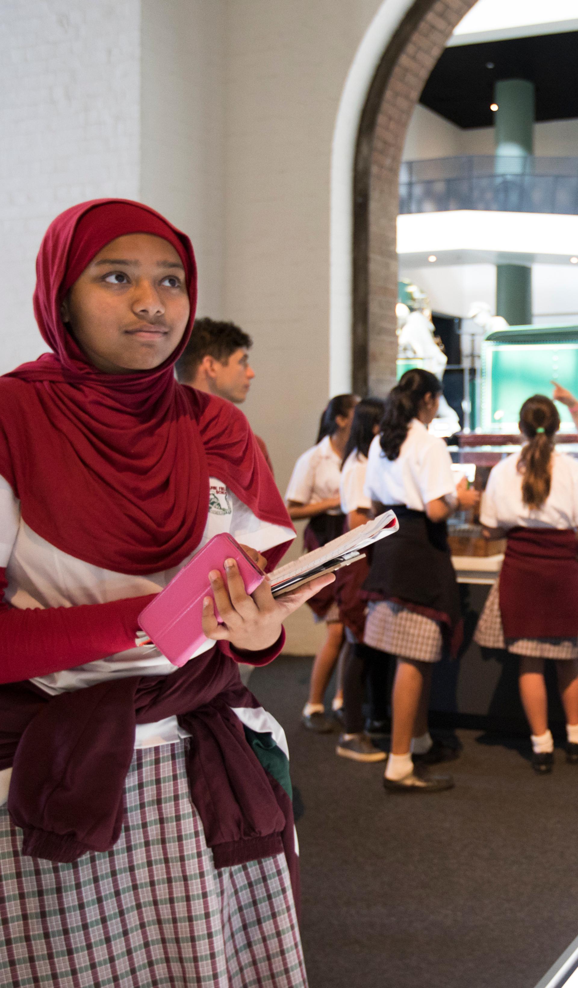young people in the background engaged in a conversation with an educator. In the foreground there is a young person looking closely at an object