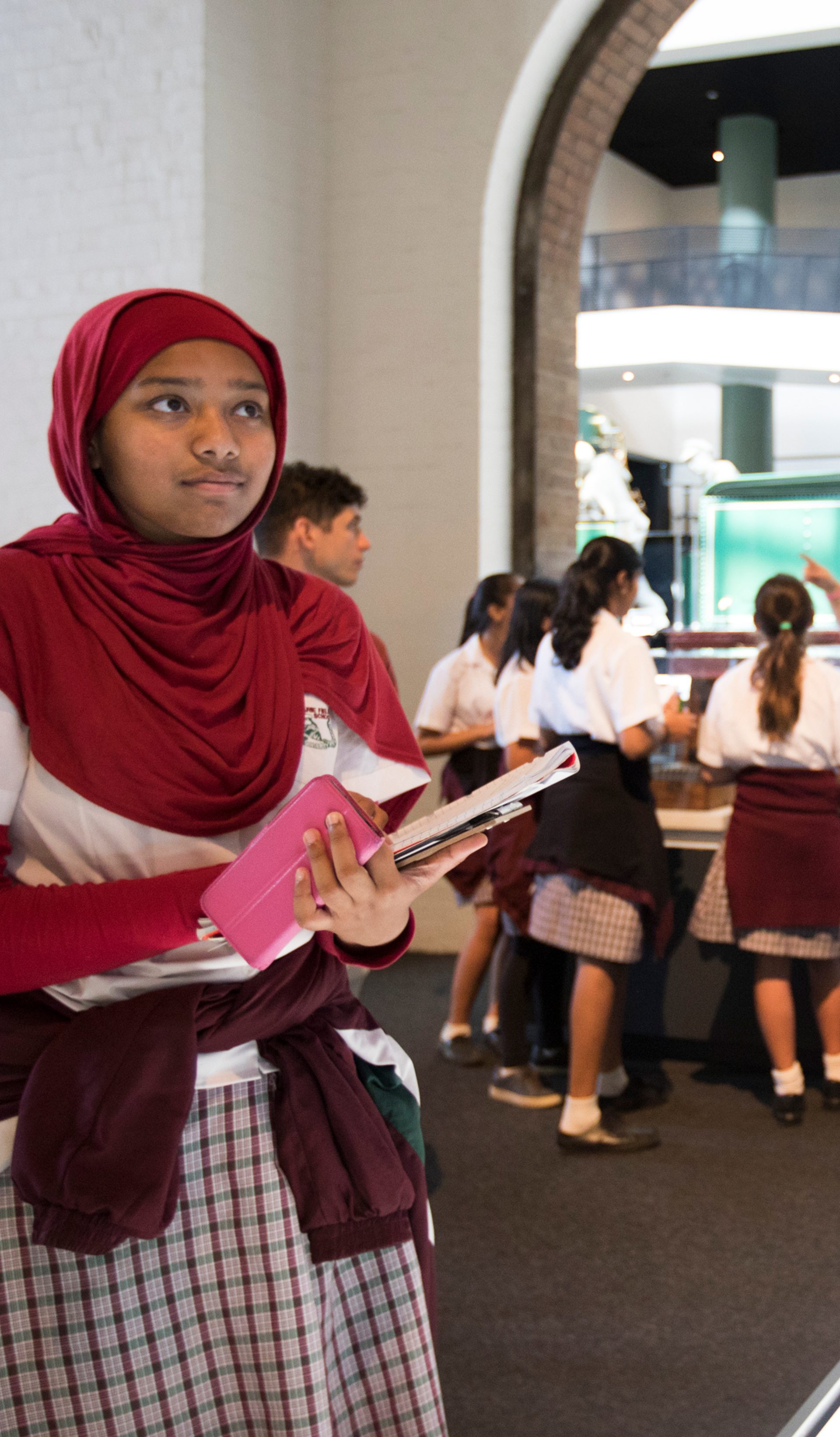 young people in the background engaged in a conversation with an educator. In the foreground there is a young person looking closely at an object
