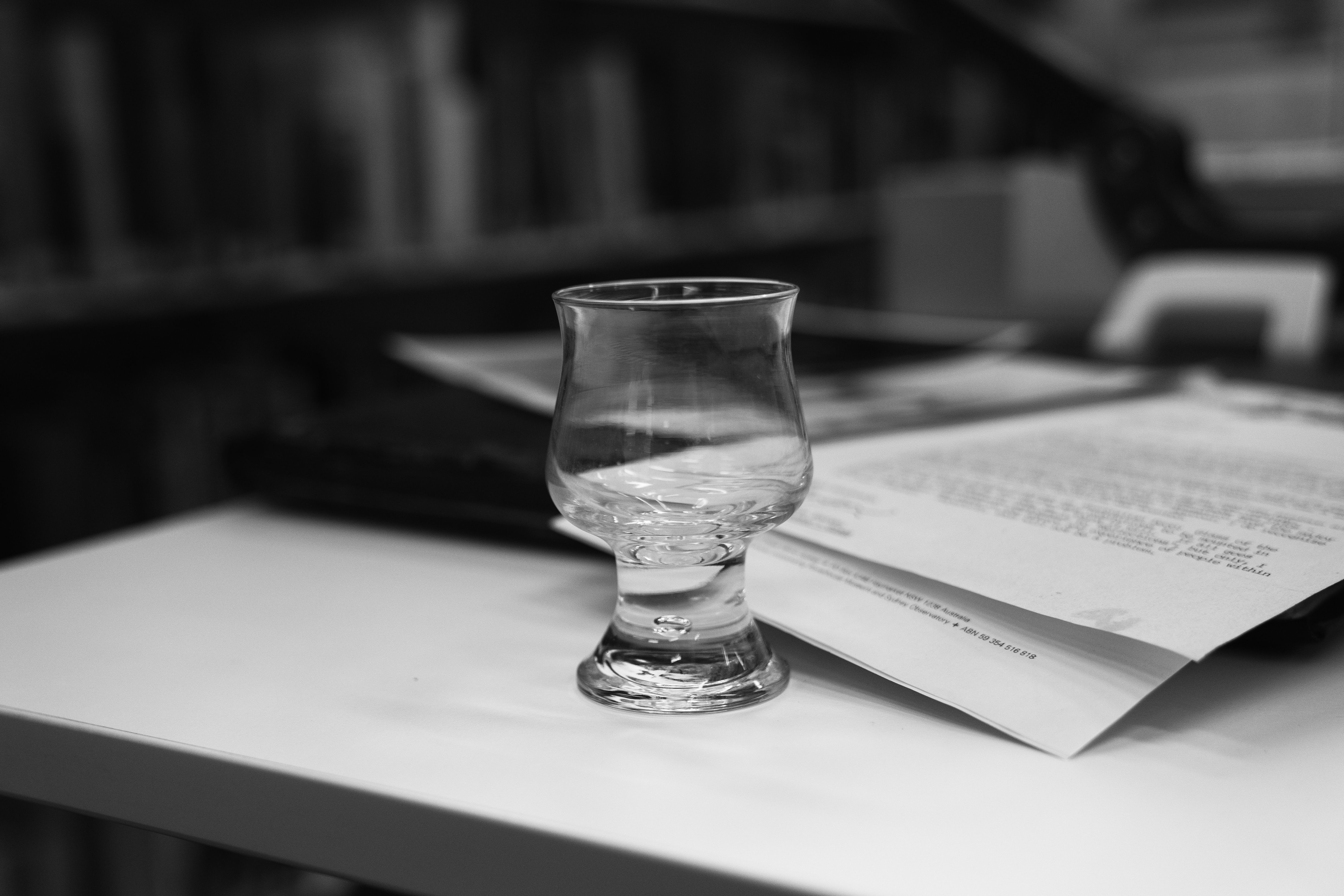 Black and white photo of a short curved glass on a table, the background shows the blurred library shelves and documents on the table.