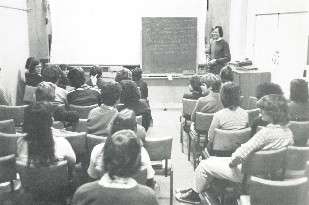 Students seated in front of teacher and chalkboard in black and white photograph