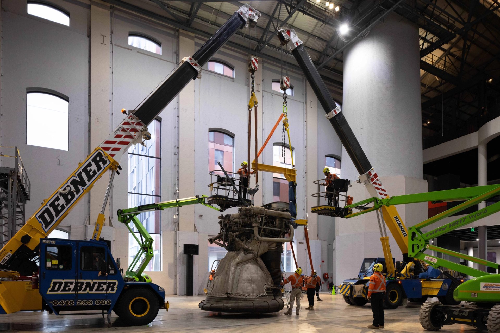 Rocket engine three times taller than average human being hoisted by cranes guided by workers wearing hi-vis.