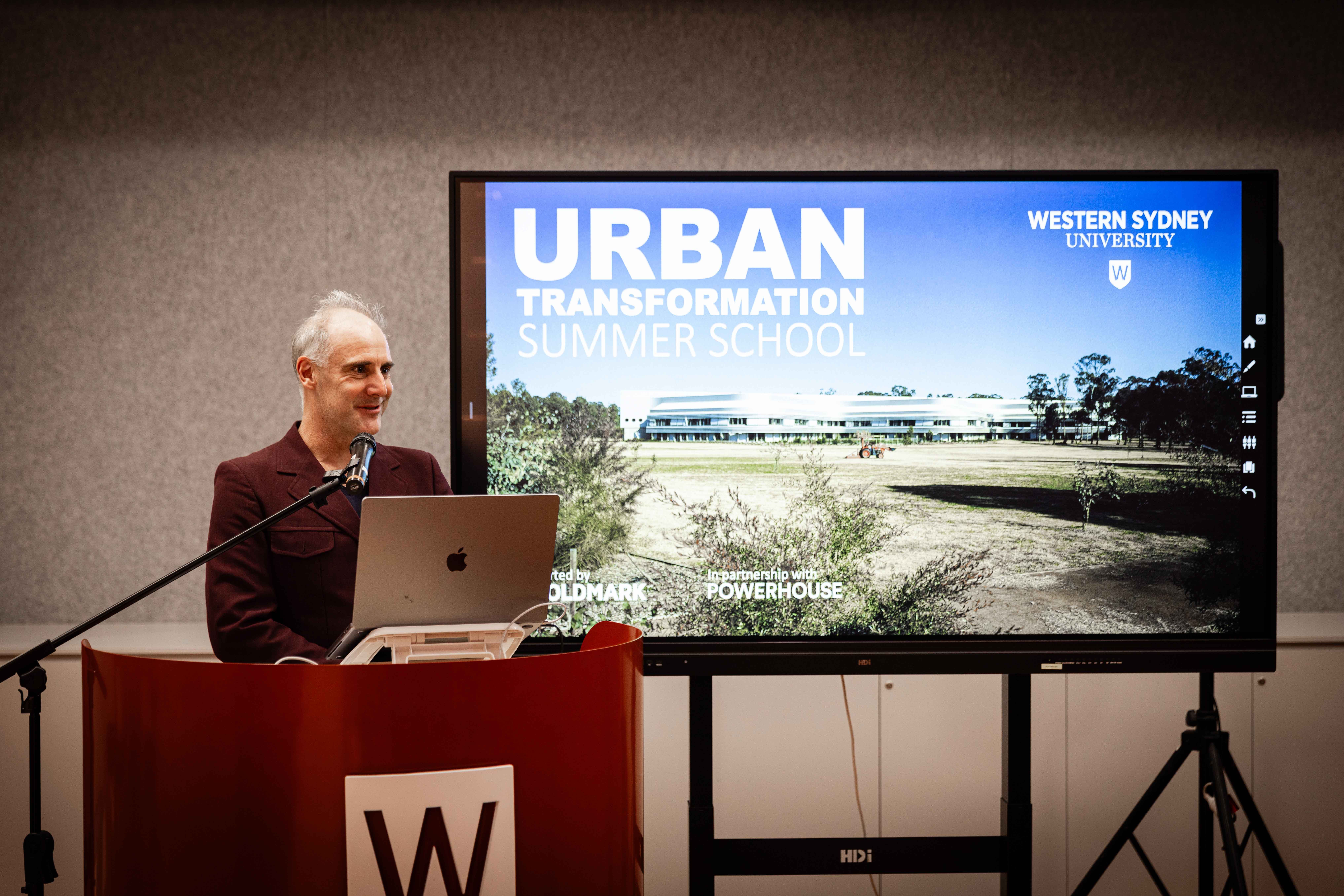 Figure standing at a lectern, presenting on a screen.