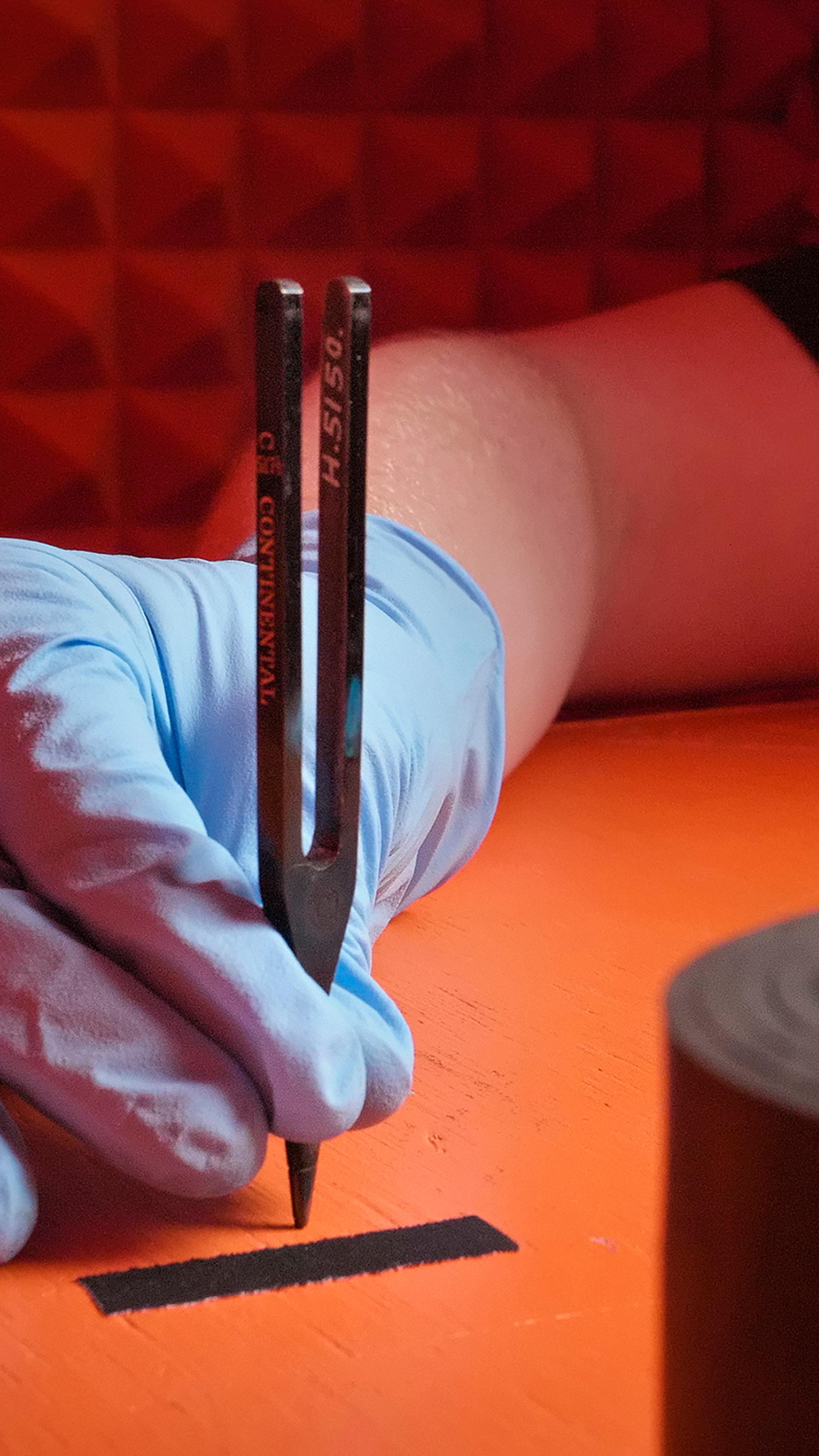 A figure wearing blue plastic gloves holds a tuning fork on an orange box.