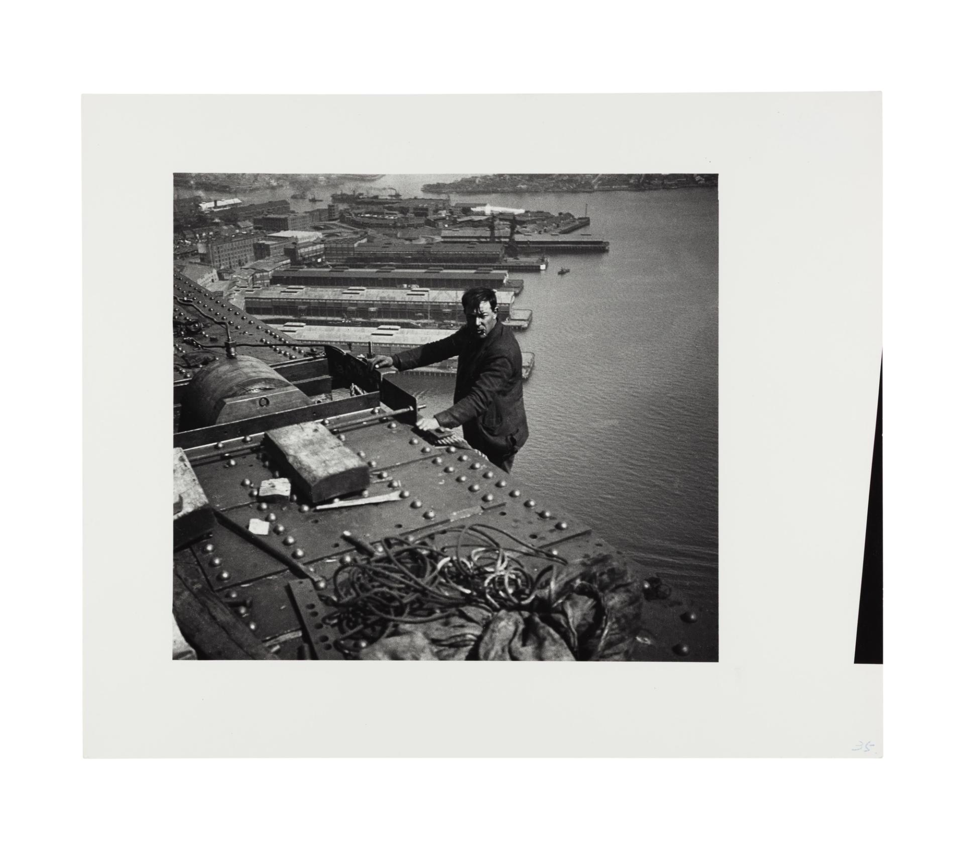 Image of a man standing on a ledge below the bridge girder visible in the photograph. He leans with both hands against the beam, smoking, and faces the camera side on. There are some construction materials on the top of the beam, rope or possible wire. The walsh bay wharves are visible in the background behind the standing figure.
