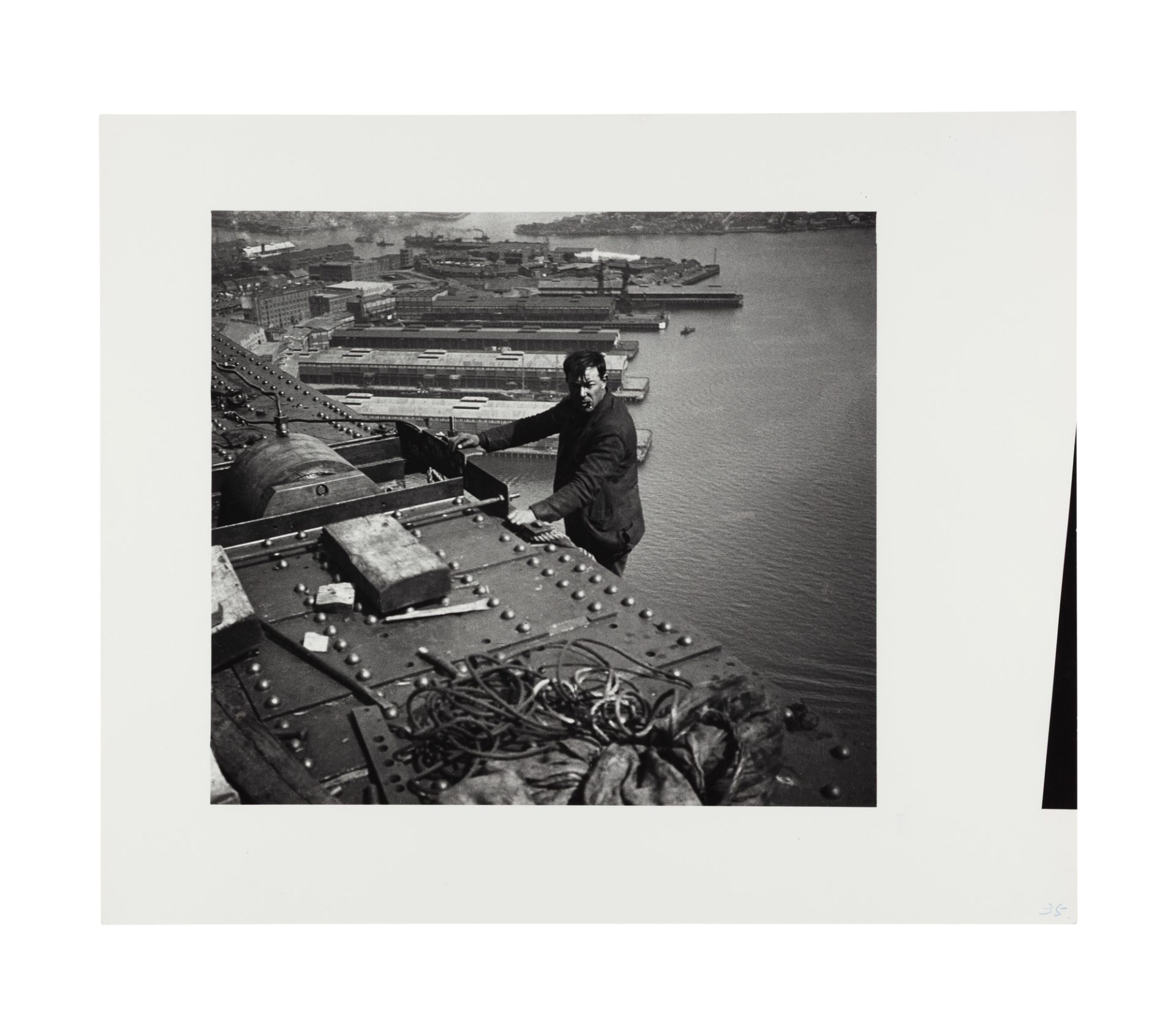 Image of a man standing on a ledge below the bridge girder visible in the photograph. He leans with both hands against the beam, smoking, and faces the camera side on. There are some construction materials on the top of the beam, rope or possible wire. The walsh bay wharves are visible in the background behind the standing figure.