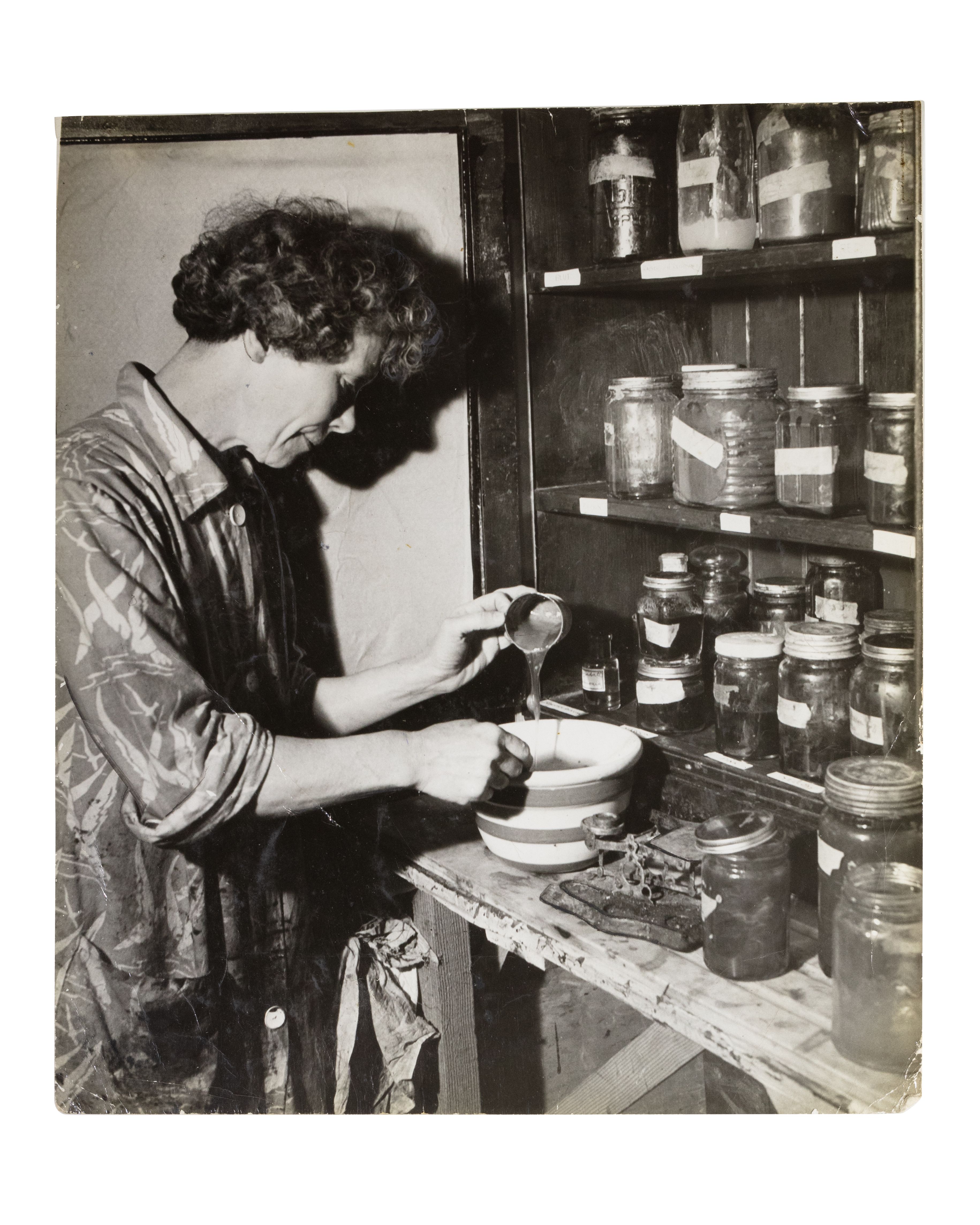 Black and white photograph depicting a person (Anne Outlaw) mixing dye in a bowl on a bench top, shelves of labelled jars in front of them.