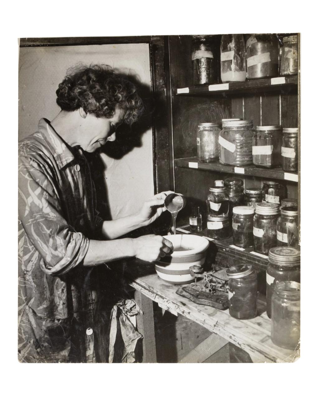 Black and white photograph depicting a person (Anne Outlaw) mixing dye in a bowl on a bench top, shelves of labelled jars in front of them.